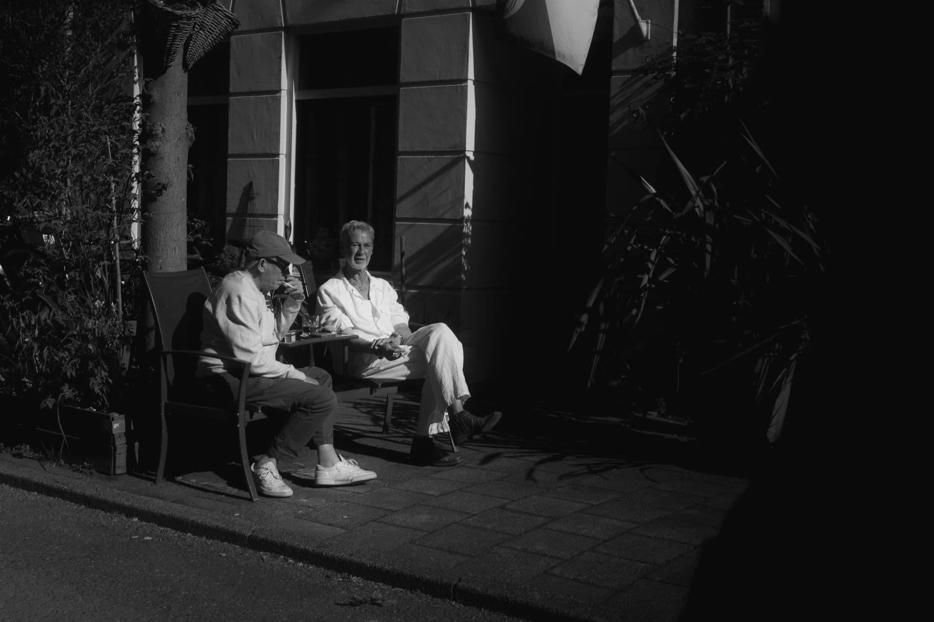Due persone siedono a un tavolino al sole, in una fotografia in bianco e nero.