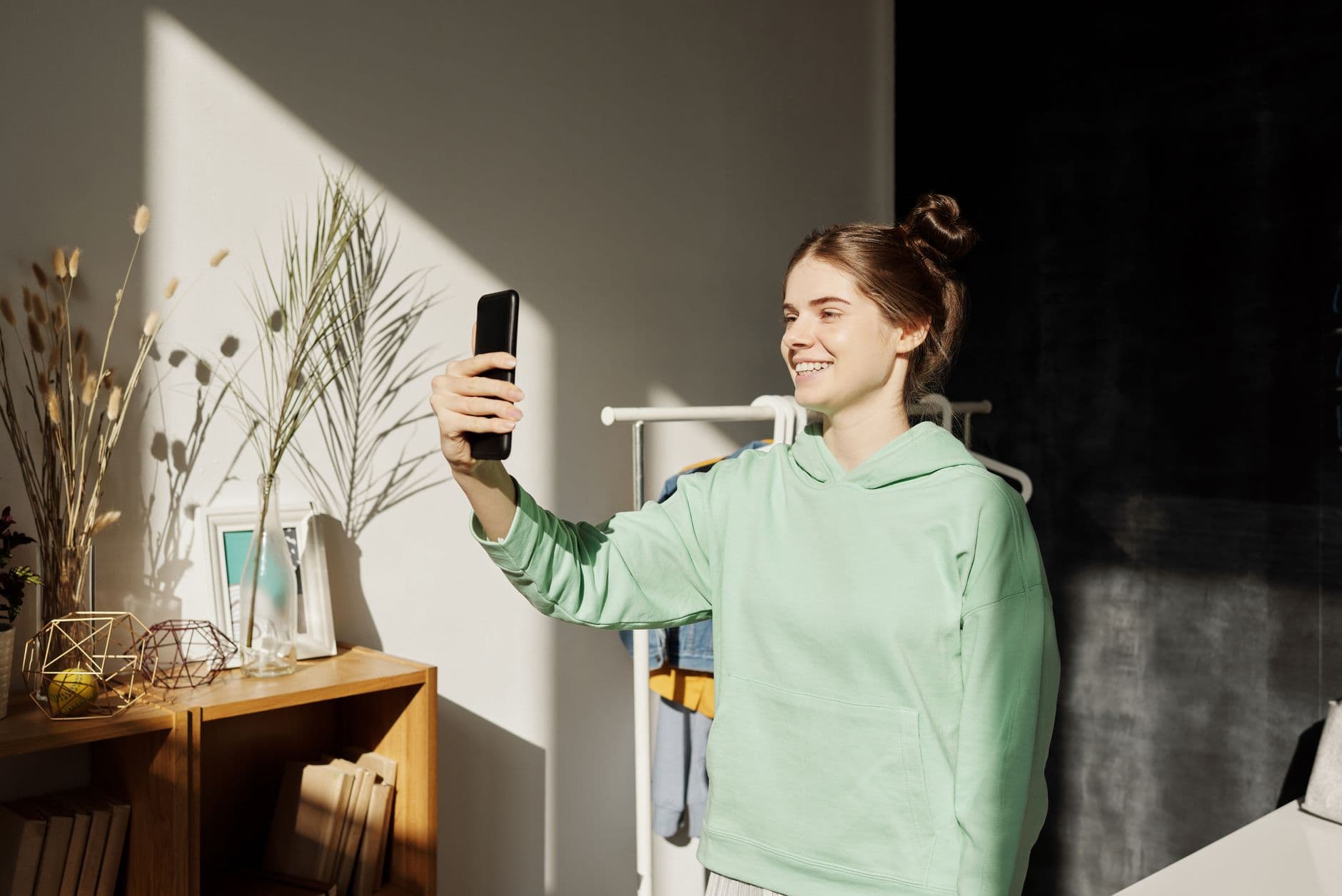 Una giovane donna sorride e si fa un selfie con il telefono in una stanza soleggiata.