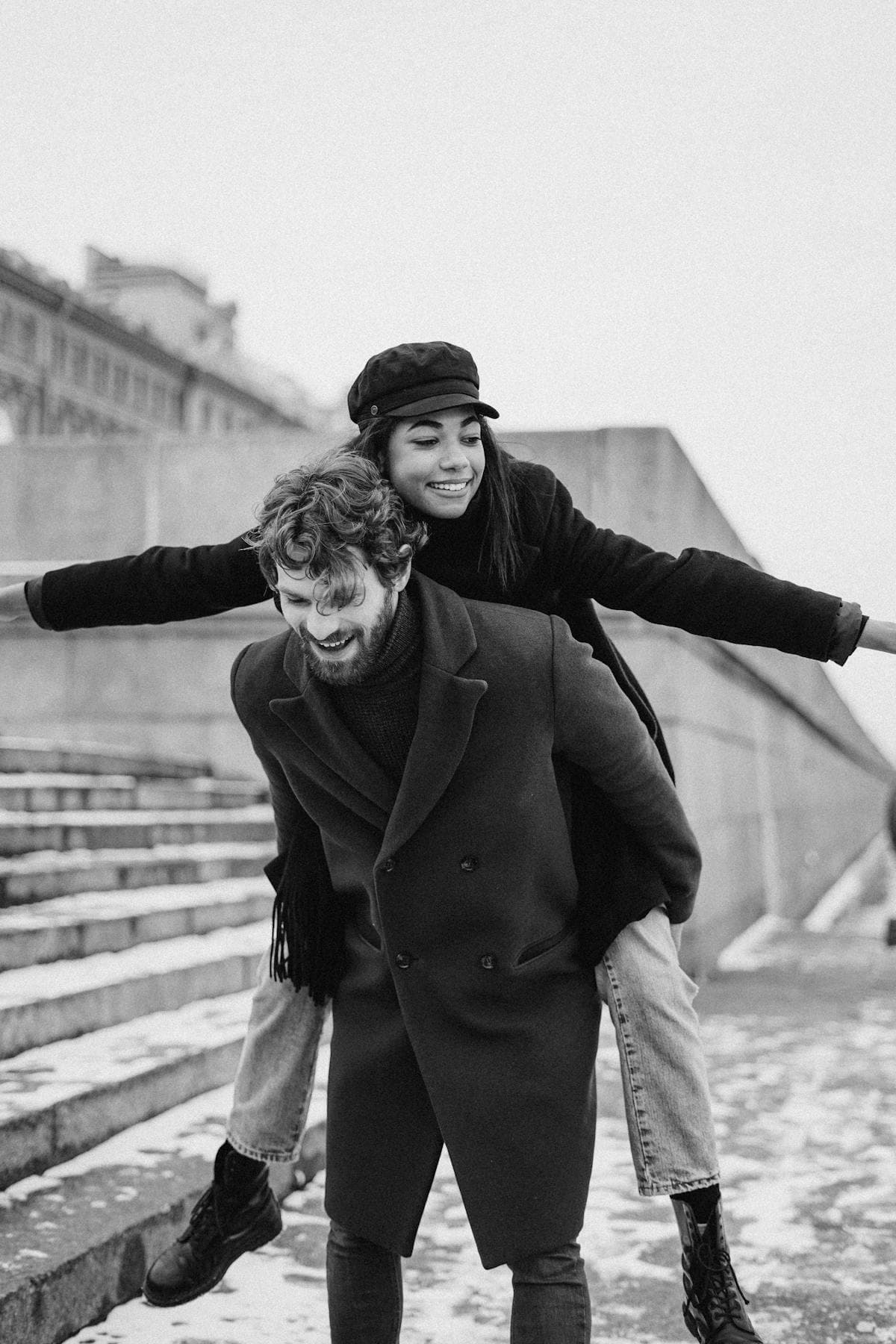 A man in a dark coat gives a woman in a black cap a piggyback beside stone steps, both laughing.