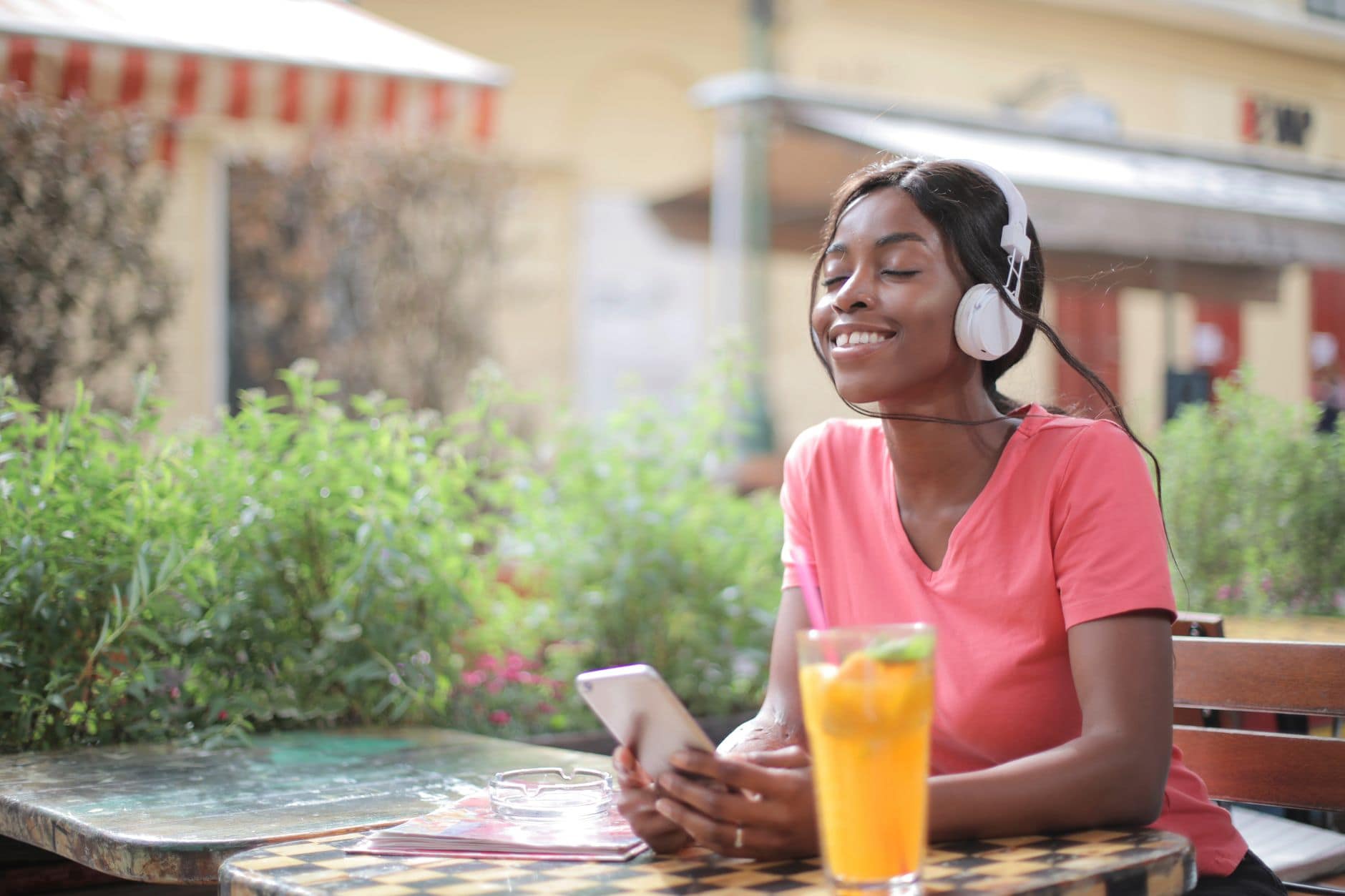 Una mujer adulta sonríe con auriculares y un celular en una mesa de café al aire libre.