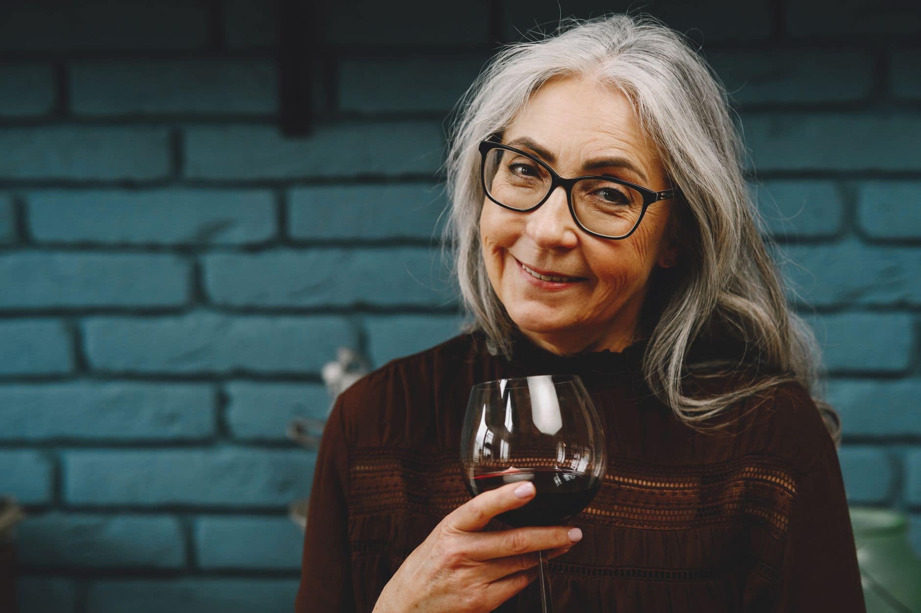 Una mujer adulta de pelo gris y anteojos sonríe mientras sostiene una copa de vino.