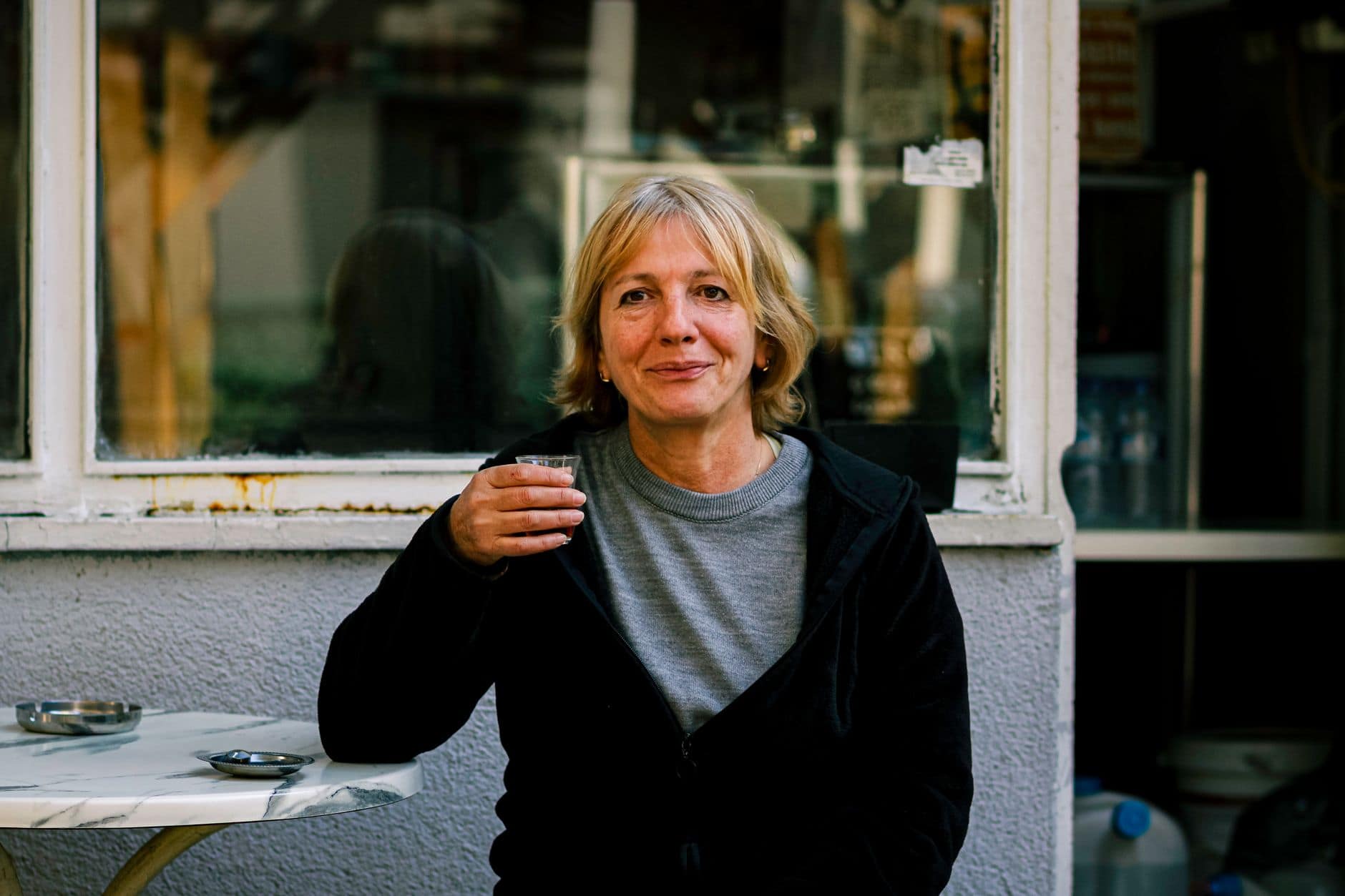 Una mujer adulta sostiene un vaso pequeño y sonríe junto a la ventana de un café.
