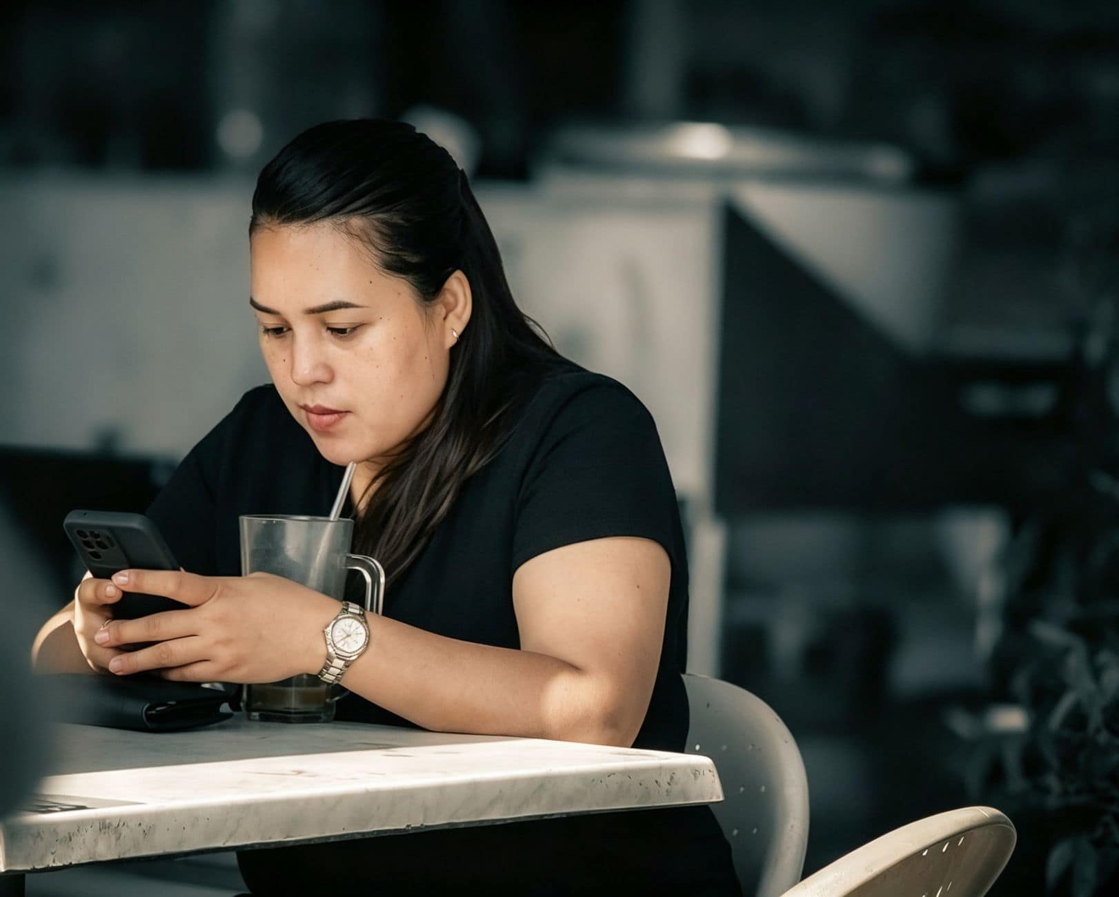 O femeie în tricou negru privește telefonul lângă o băutură rece, la o masă de cafenea