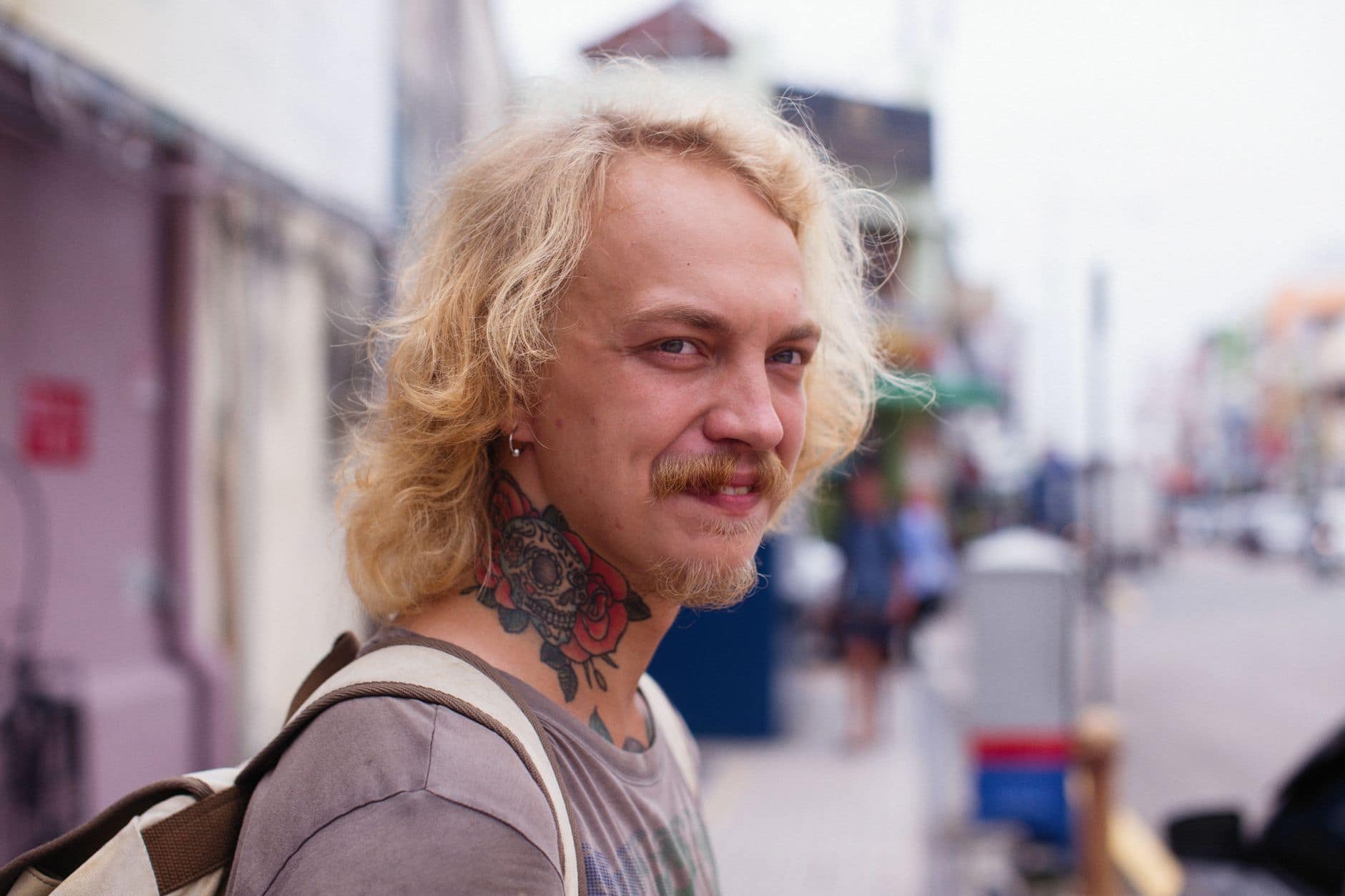 Homem adulto loiro com bigode e tatuagem no pescoço sorri em uma rua movimentada.
