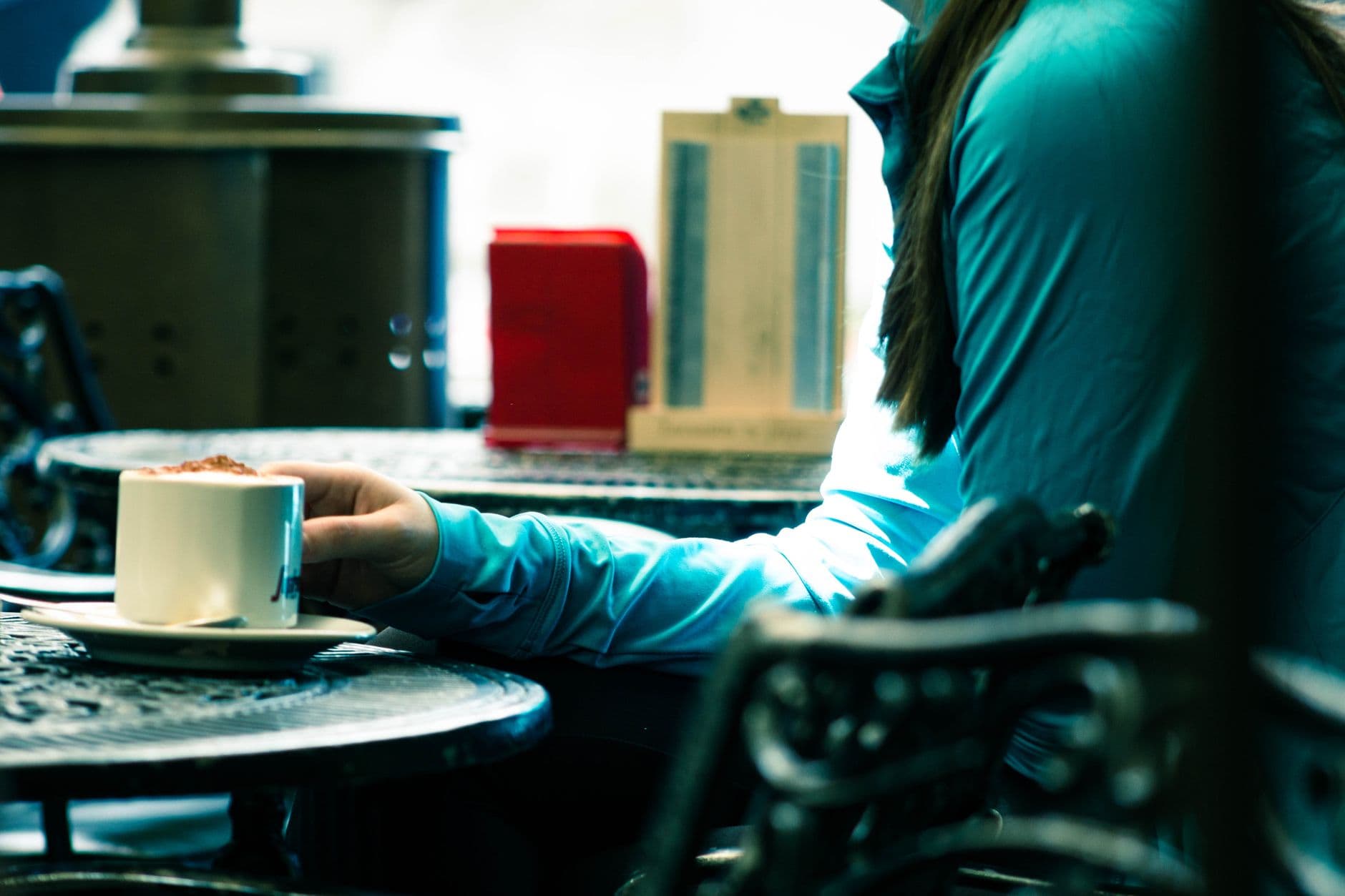 Iemand in een lichtblauwe trui houdt een kop koffie vast aan een tafeltje in een café.