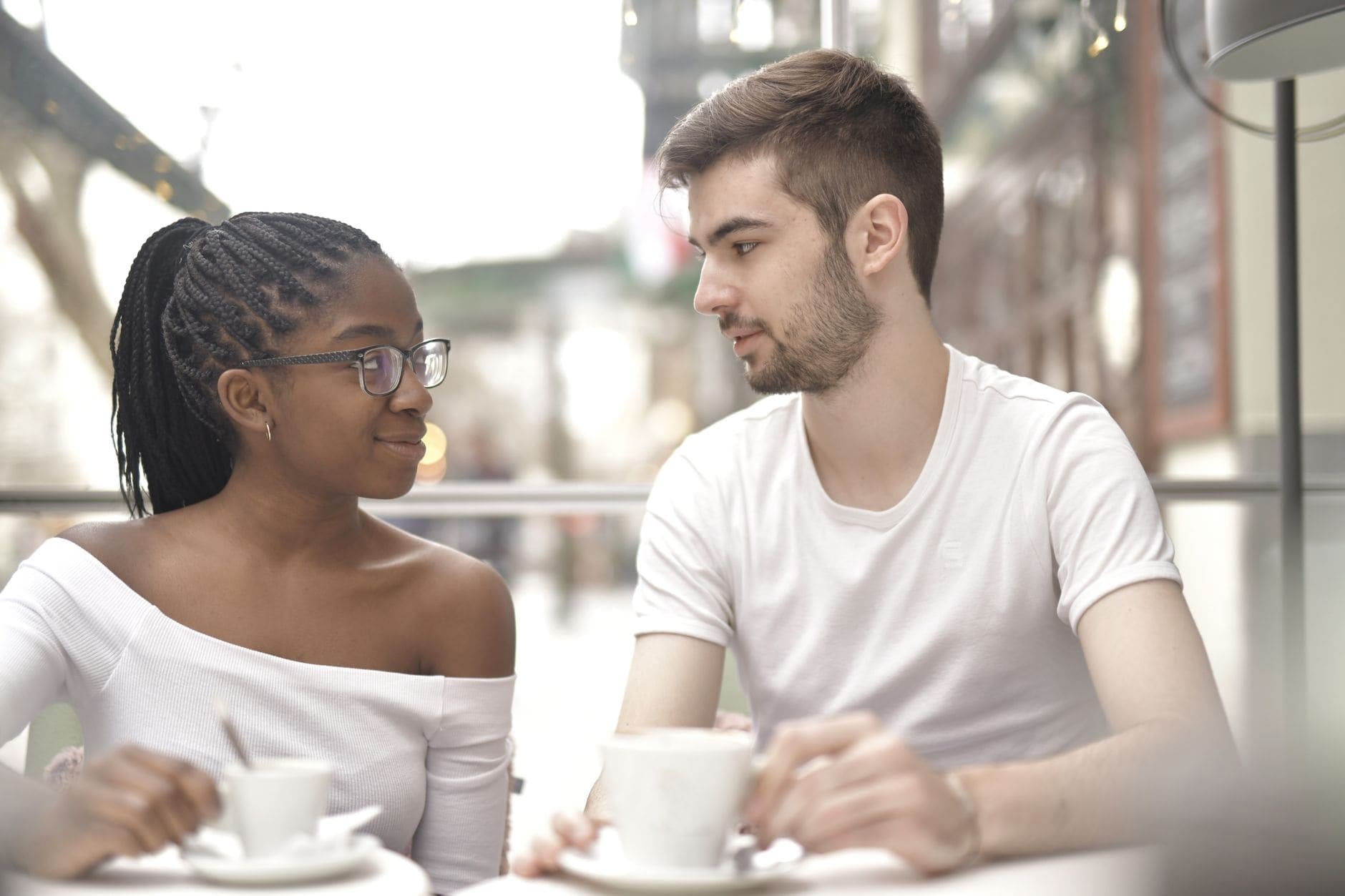 Uma mulher de óculos e um homem conversam com chávenas numa mesa junto à janela.