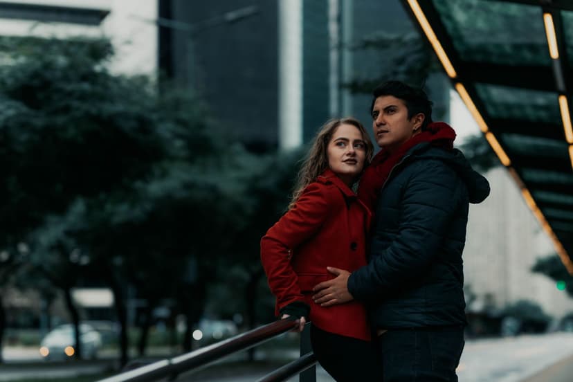 A couple in warm winter coats stand close together beside a railing on an overcast city street, the woman in a bright red coat leaning into her partner as office towers rise behind them.