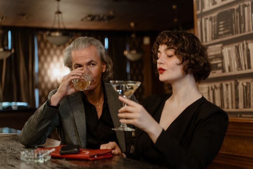 A silver-haired man in a grey blazer sips whisky beside a younger woman in a black dress holding a martini glass at the counter of a dimly lit, wood-panelled bar.