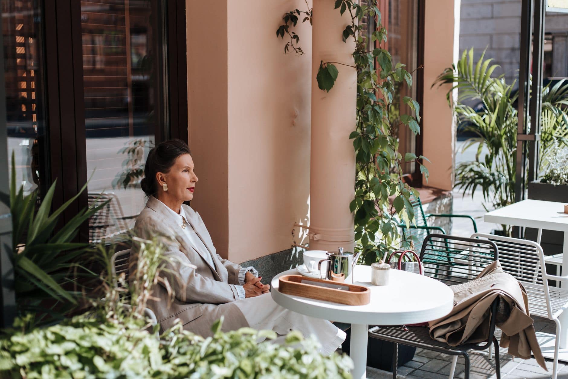O femeie în costum crem stă la o masă albă cu ceainic și ceașcă, între plantele unei cafenele