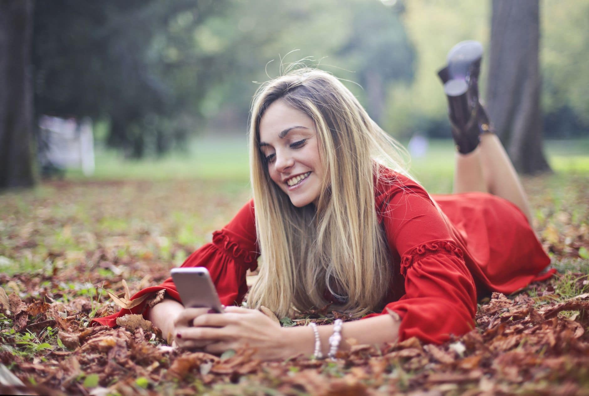 Een vrouw in een rode jurk ligt tussen de bladeren en glimlacht naar haar telefoon.