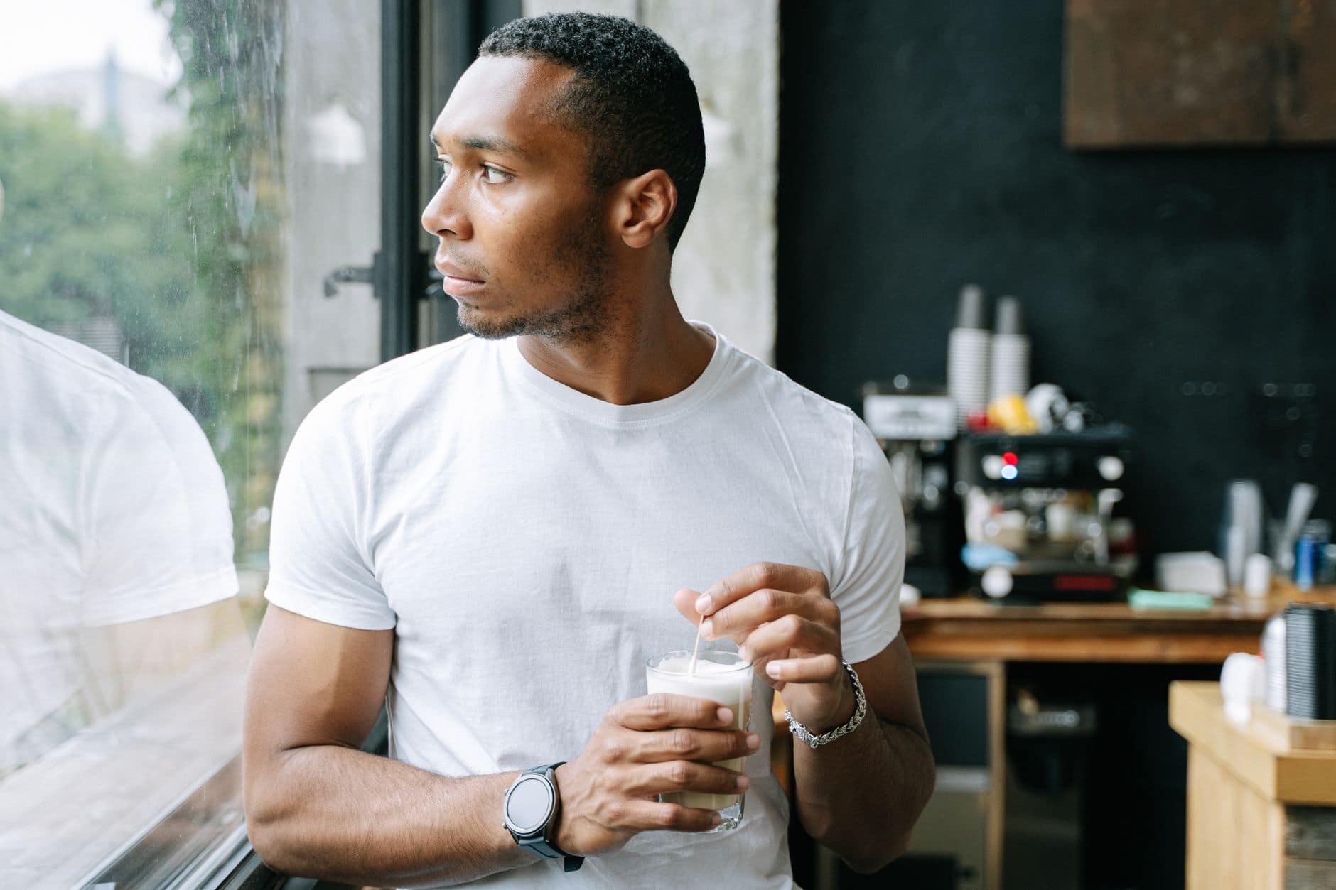 Een man in een wit T-shirt houdt een glas vast en kijkt uit het raam van een café.