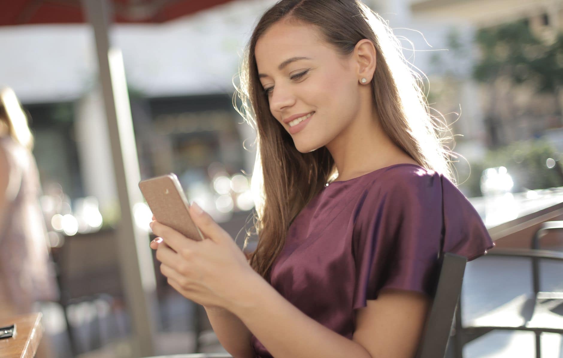 Een vrouw in een paarse blouse glimlacht naar haar telefoon aan een cafétafel.