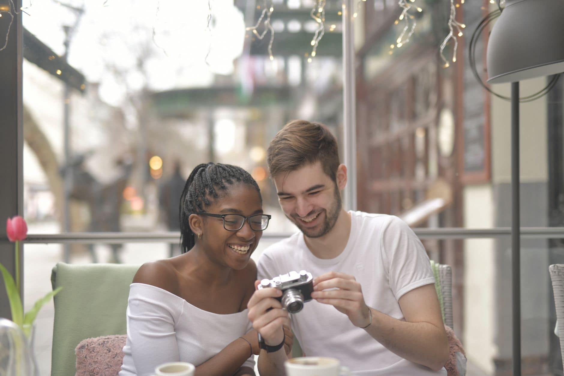 Een vrouw en een man bekijken glimlachend een fototoestel bij het raam van een café.