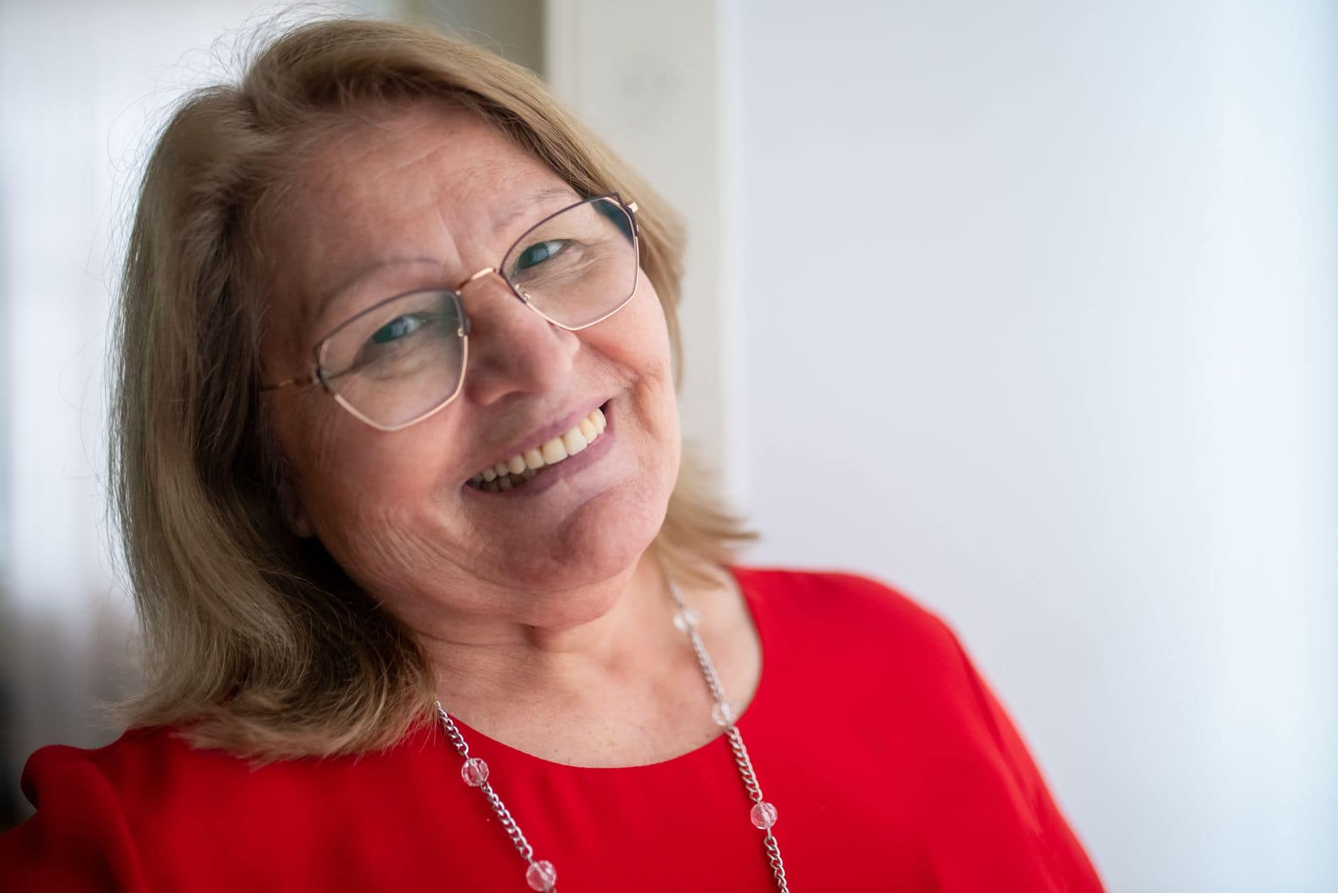 Porträt einer lächelnden Frau mit Brille und rotem Oberteil am Fenster.
