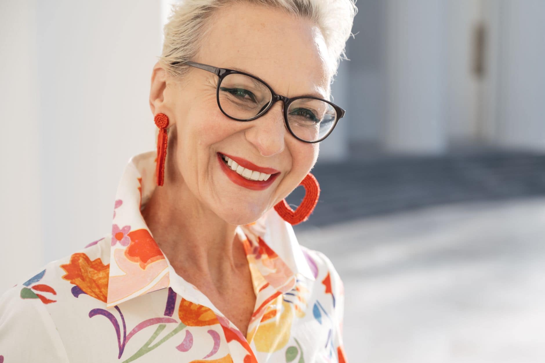 Eine Frau mit Brille, roten Ohrringen und geblümter Bluse lächelt vor einem hellen Hintergrund.