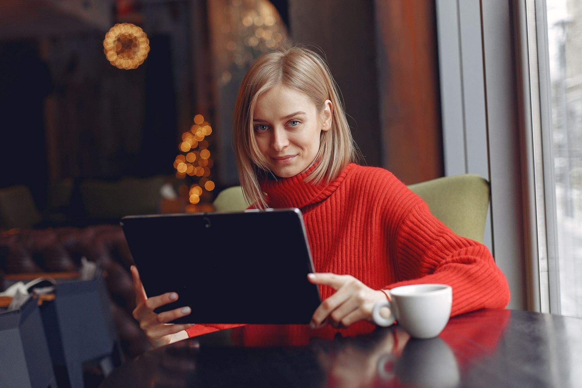 A woman in a red roll-neck sweater holds a tablet at a café table beside a white cup.