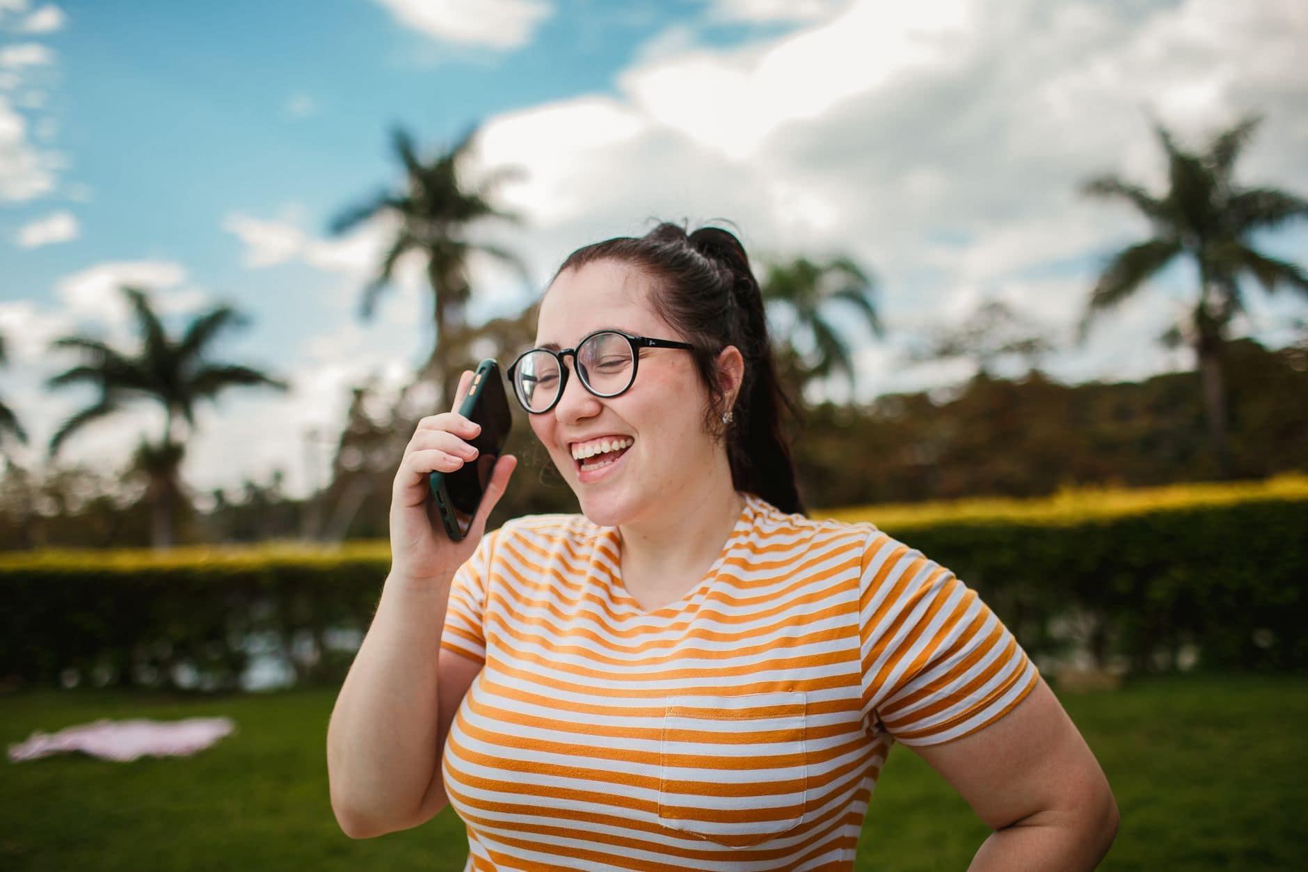 Eine Frau mit Brille und gestreiftem Shirt lacht beim Telefonieren im Freien.