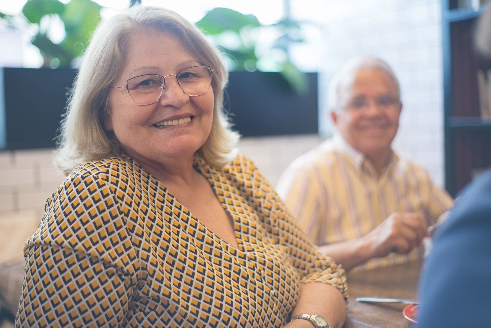A woman with glasses and a patterned blouse smiles, with a man seated behind her.