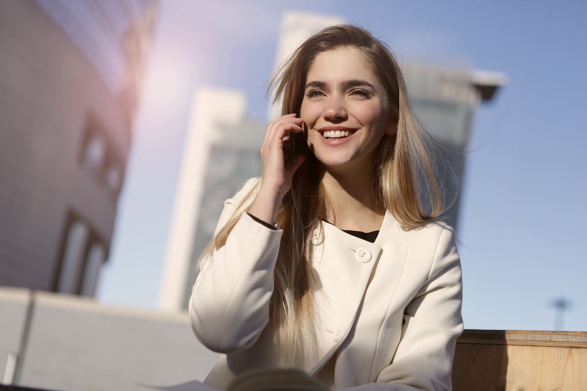 A smiling woman in a white coat takes a phone call outdoors in bright sunlight beside city buildings