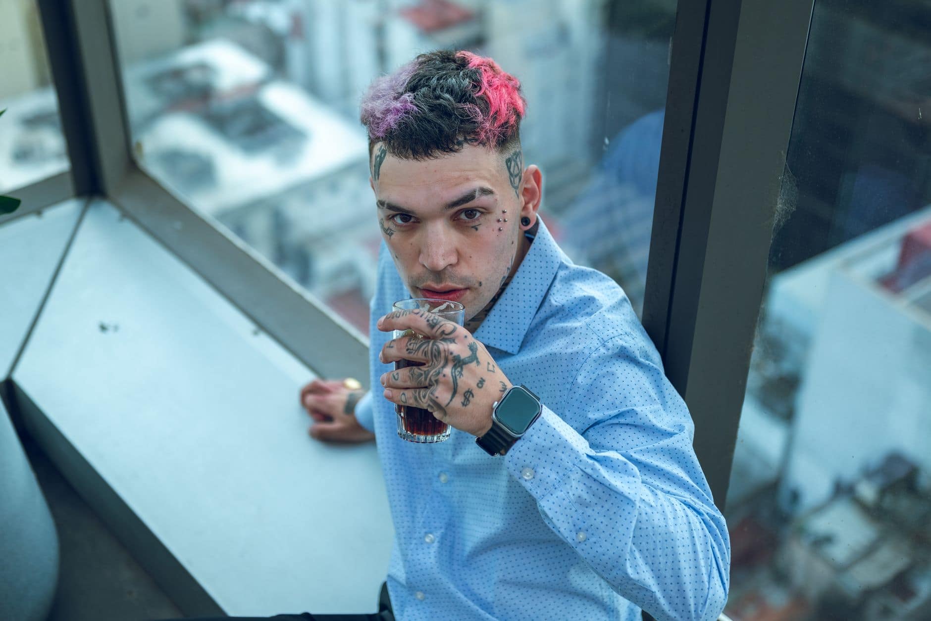 Ein Mann im hellblauen Hemd sitzt mit einem Glas am Fenster und schaut in die Kamera.