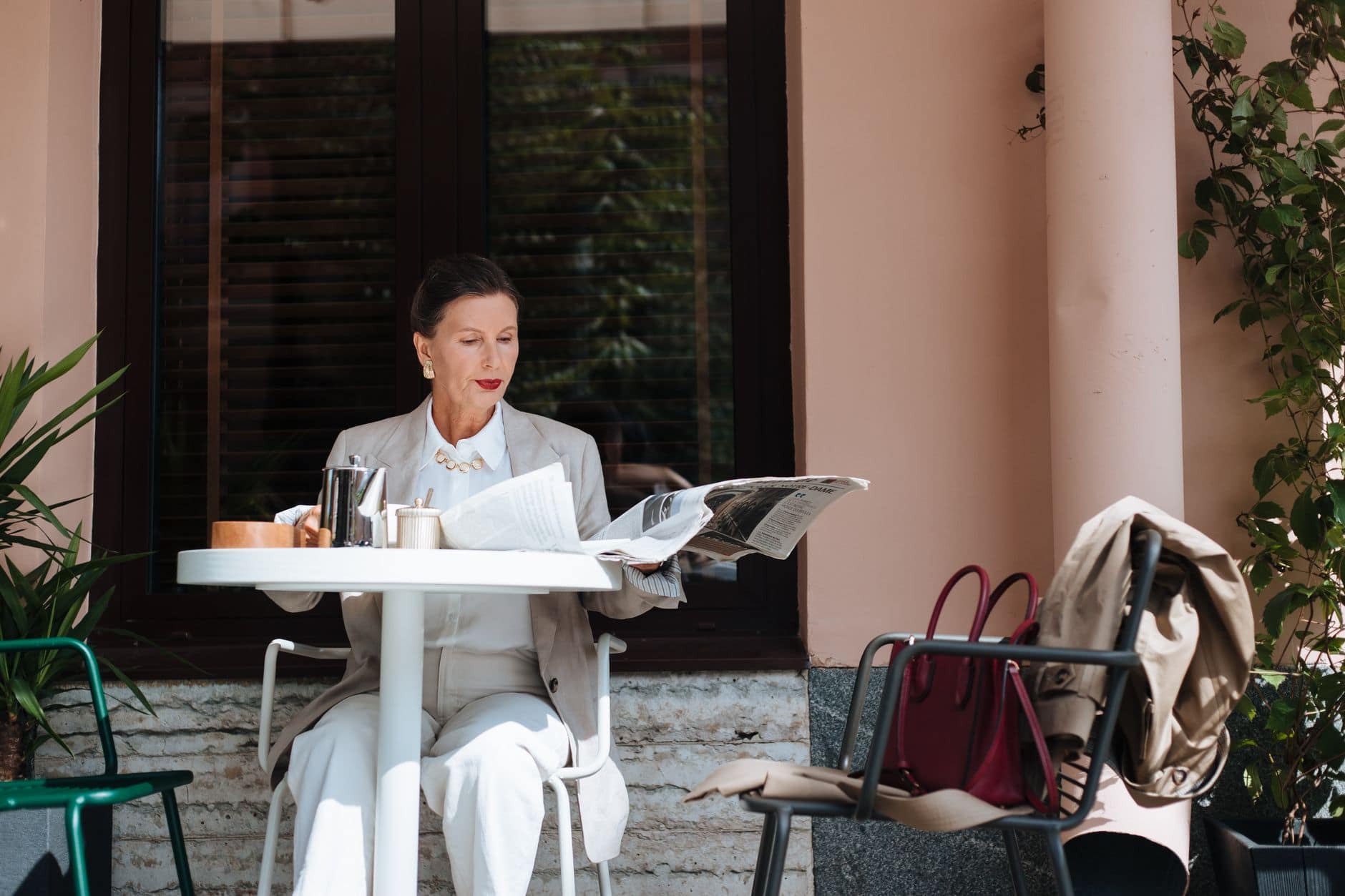 Eine Frau in einem hellen Anzug liest an einem Cafétisch im Freien eine Zeitung.