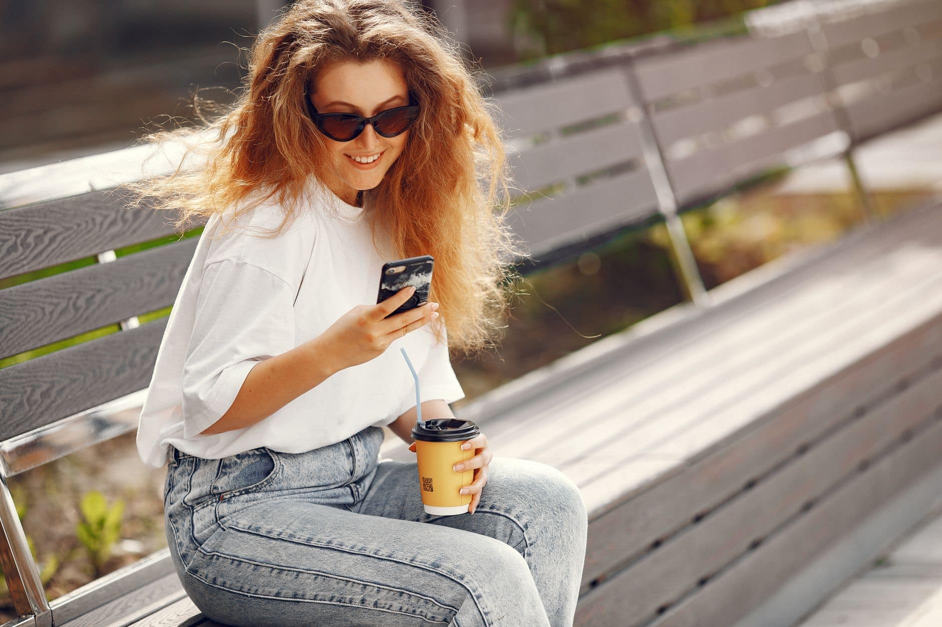 Eine Frau mit Sonnenbrille lächelt auf einer Holzbank mit Handy und gelbem Kaffeebecher in den Händen.