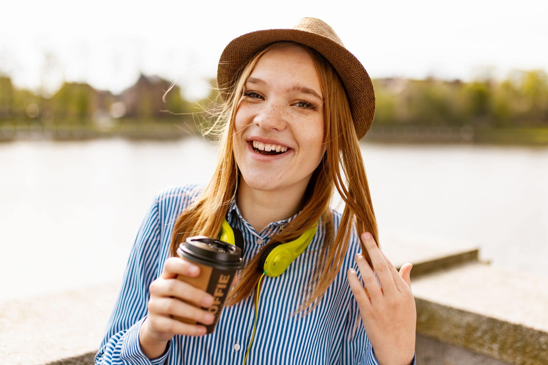 Eine lächelnde Frau mit braunem Hut und Kopfhörern hält einen Kaffeebecher am Wasser.