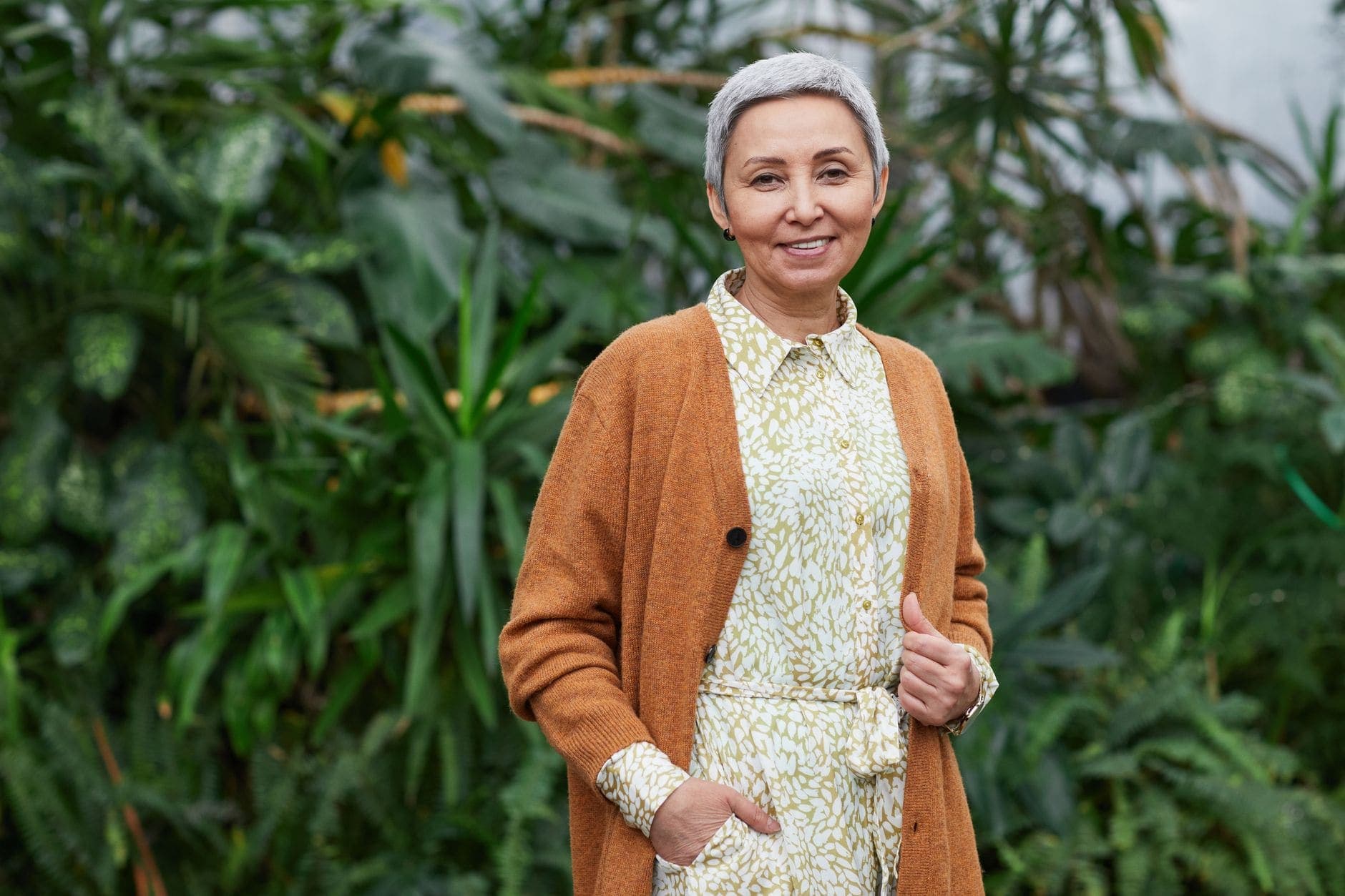 A woman with short grey hair smiles in a brown cardigan and patterned dress beside green plants.