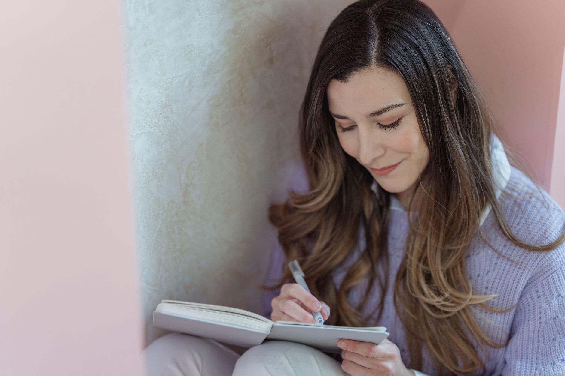 A woman in a lilac jumper writes in an open notebook beside a pale curtain.
