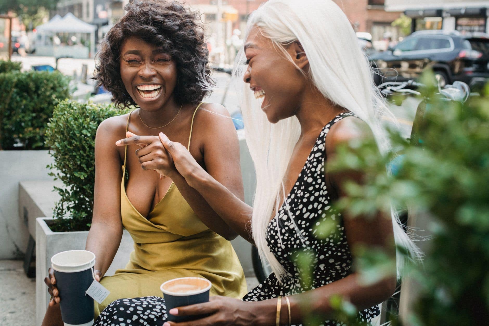 Zwei Frauen in Sommerkleidern lachen beim Kaffee an einem Tisch im Freien.