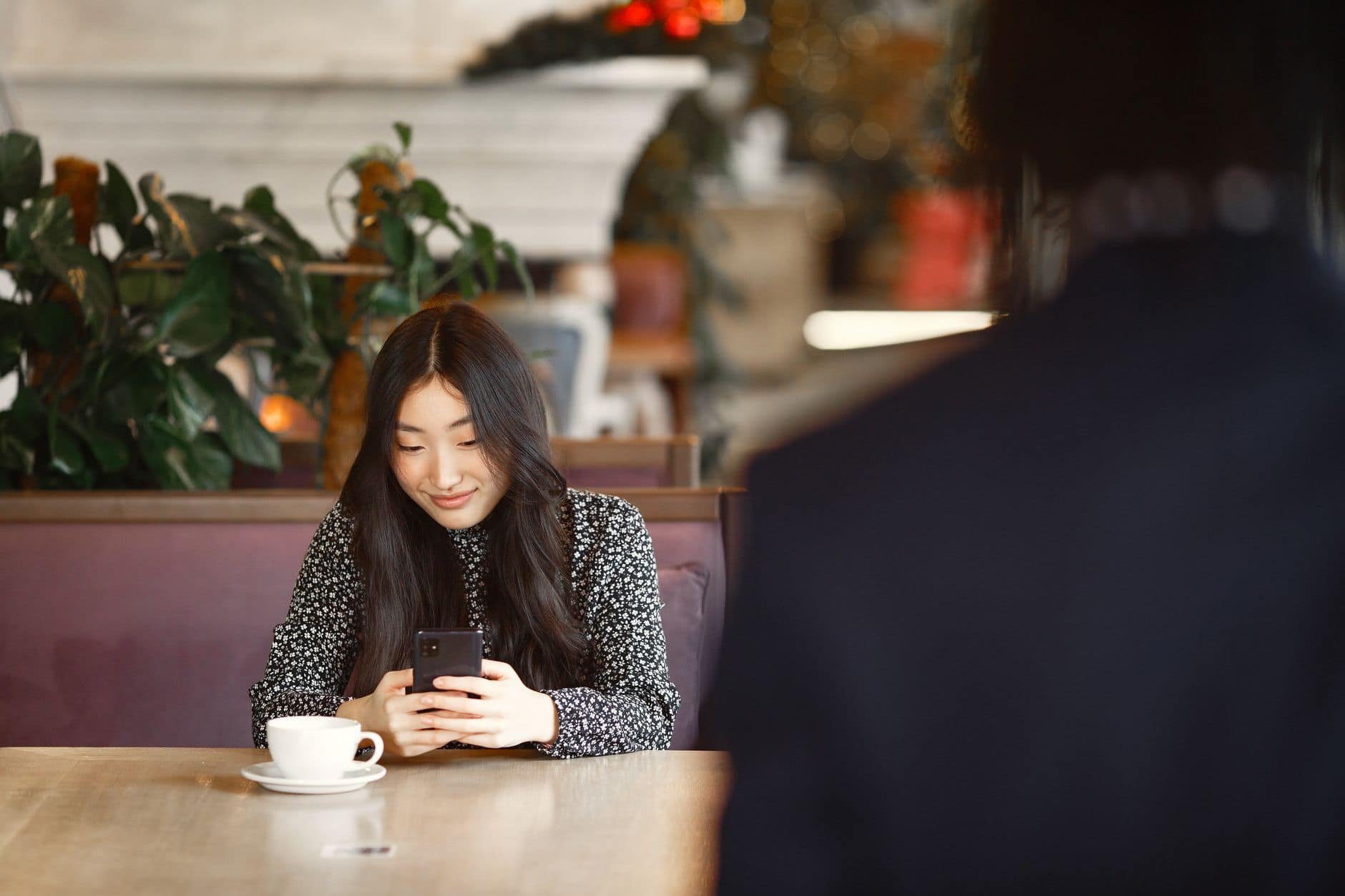 Eine Frau im geblümten Oberteil lächelt auf ihr Handy, vor ihr steht eine weiße Kaffeetasse.