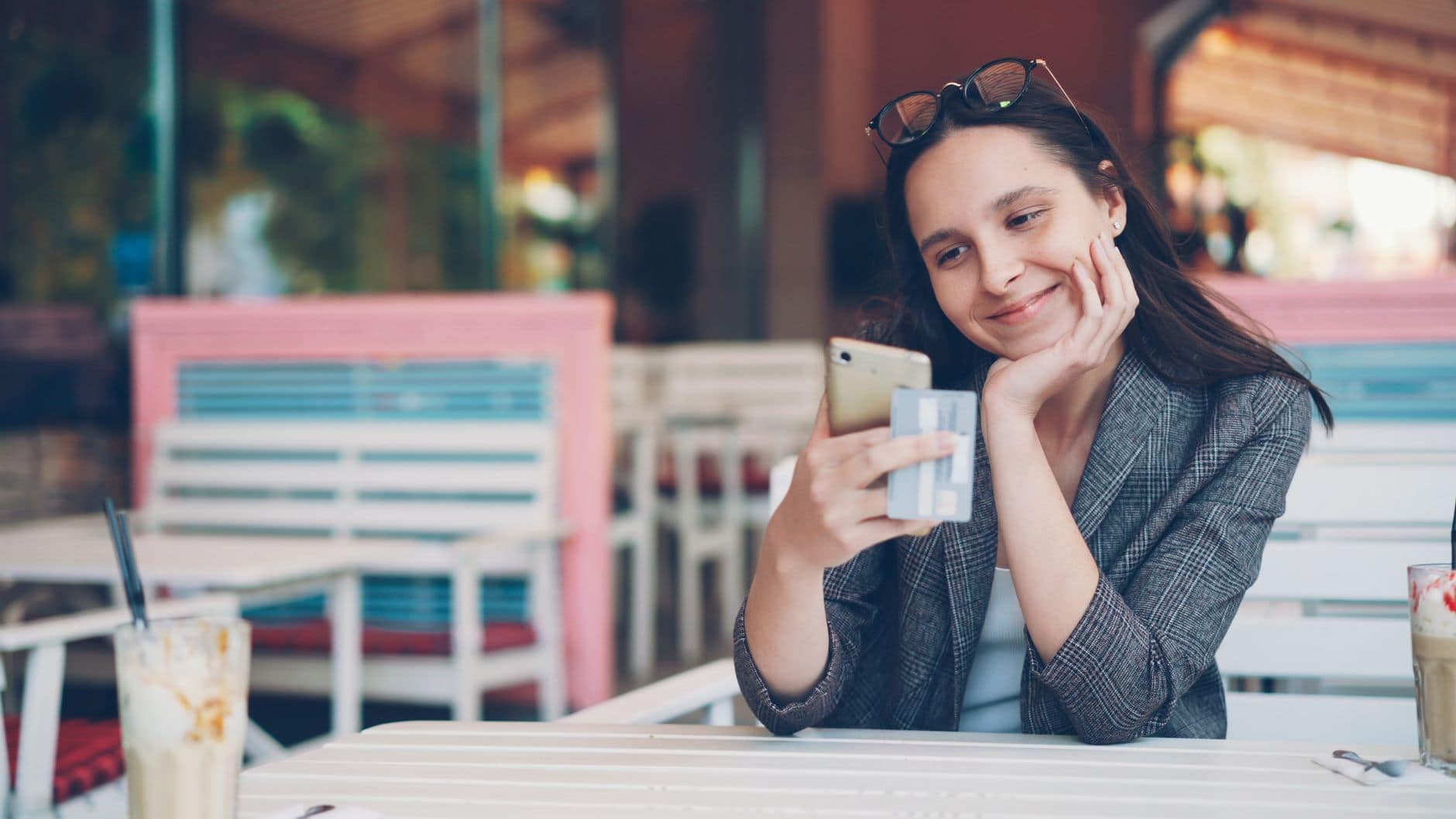 Eine Frau im grauen Blazer betrachtet ihr Smartphone und hält eine Karte am Cafétisch.