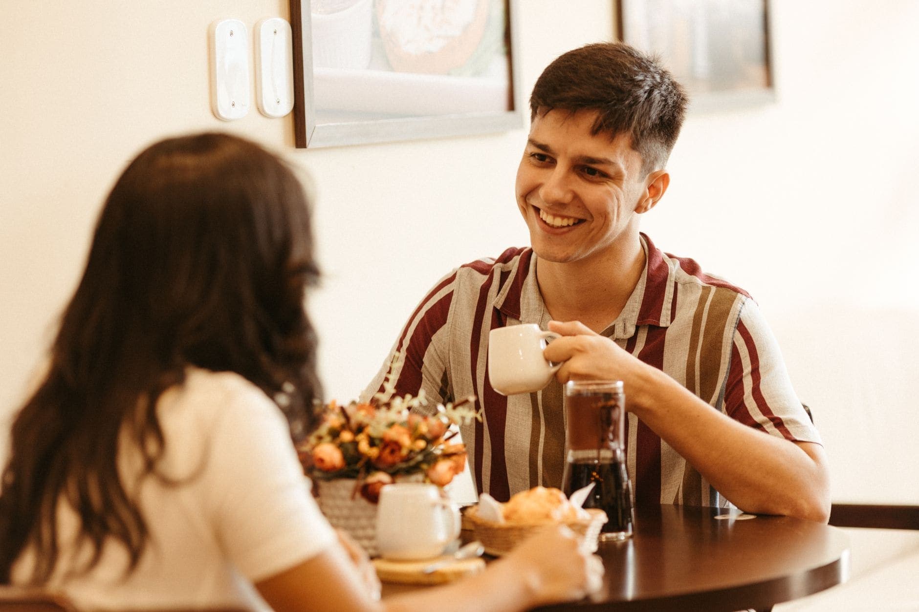 Um homem sorri com uma chávena na mão, à mesa de um café claro, frente à sua companhia
