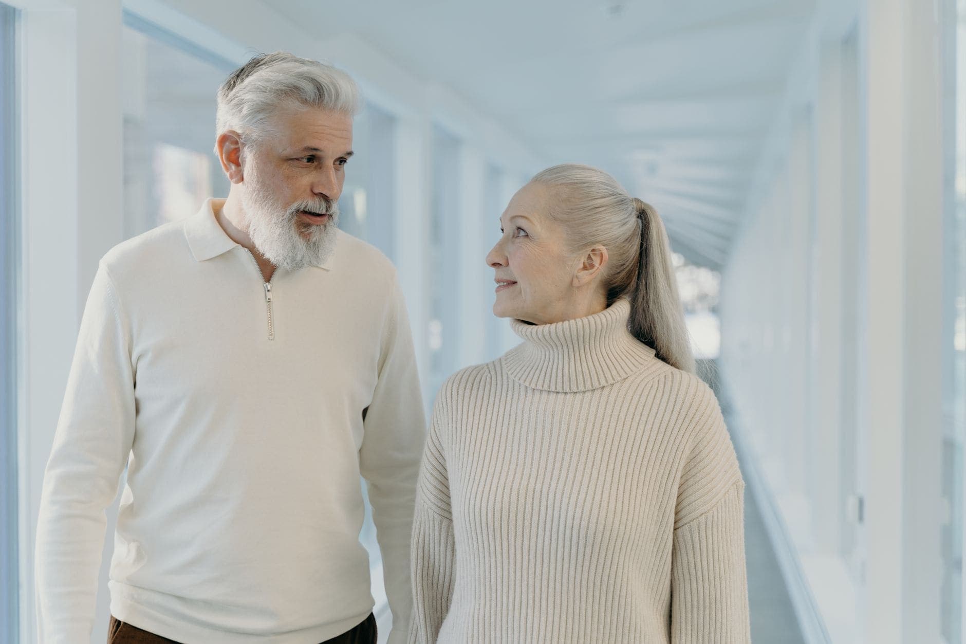 Um homem de barba branca conversa com uma mulher de camisola clara num corredor luminoso.