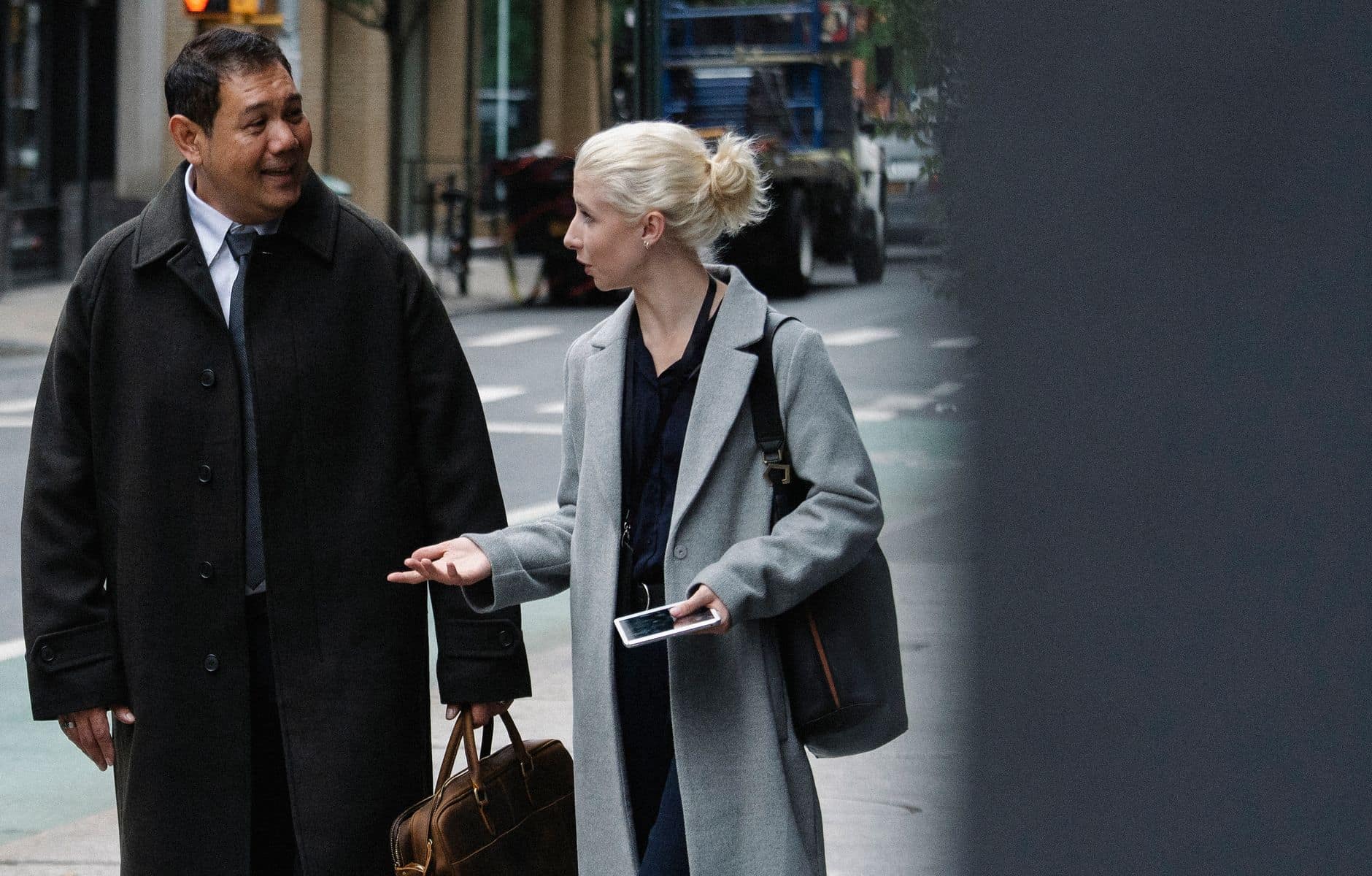 Une jeune femme blonde en manteau gris discute avec un homme d’âge mûr en pardessus dans une rue de la ville.