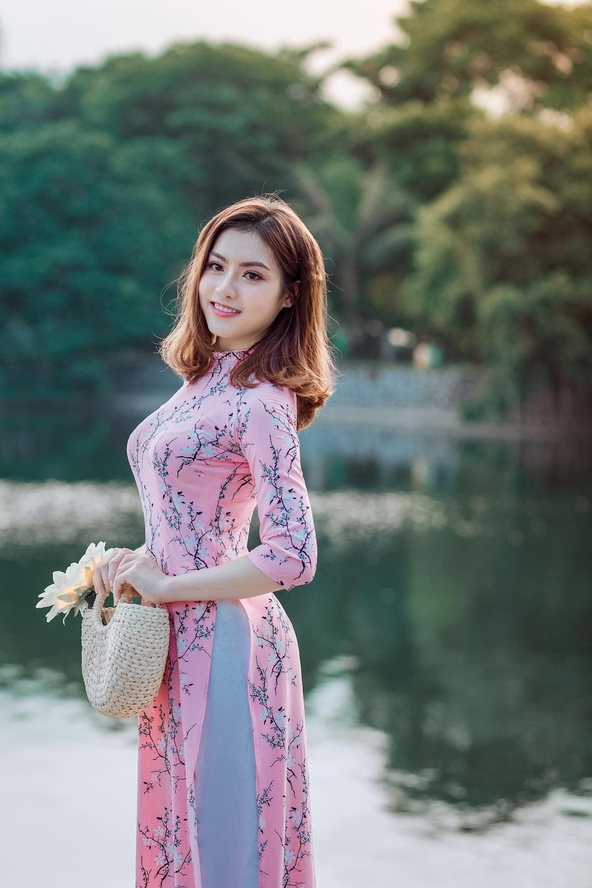 A woman in a pink floral dress holds a woven basket bag by a lake with green trees behind her.