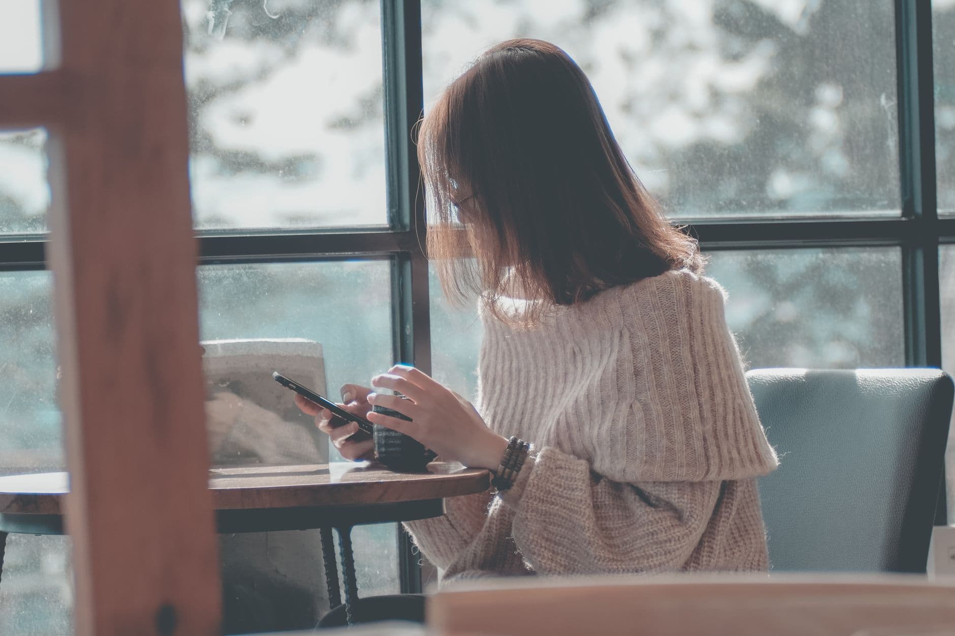 Een vrouw in een dikke crèmekleurige trui typt op haar telefoon aan een tafeltje bij het raam.