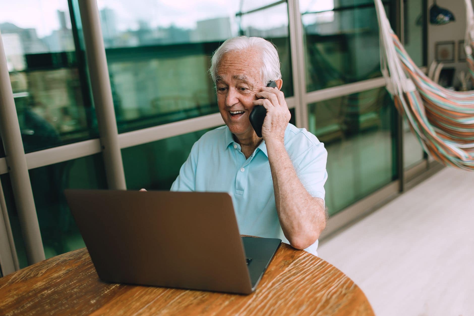 Een man in een lichtblauwe polo belt glimlachend aan een tafel met een laptop.