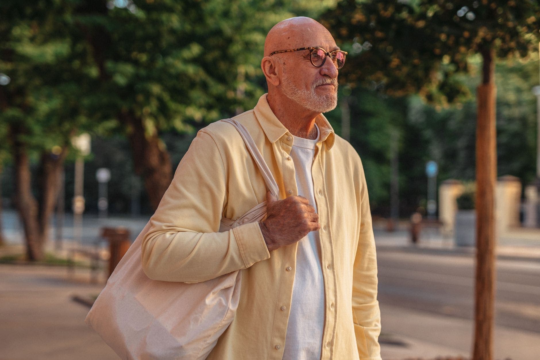 Um homem de barba branca e camisa amarela carrega uma bolsa de tecido no ombro.