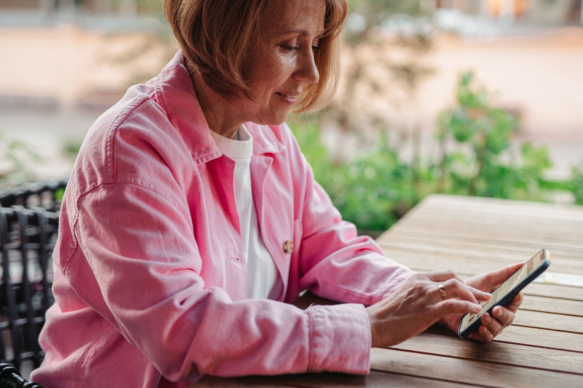 Uma mulher de camisa cor-de-rosa sorri enquanto usa o telemóvel à mesa.