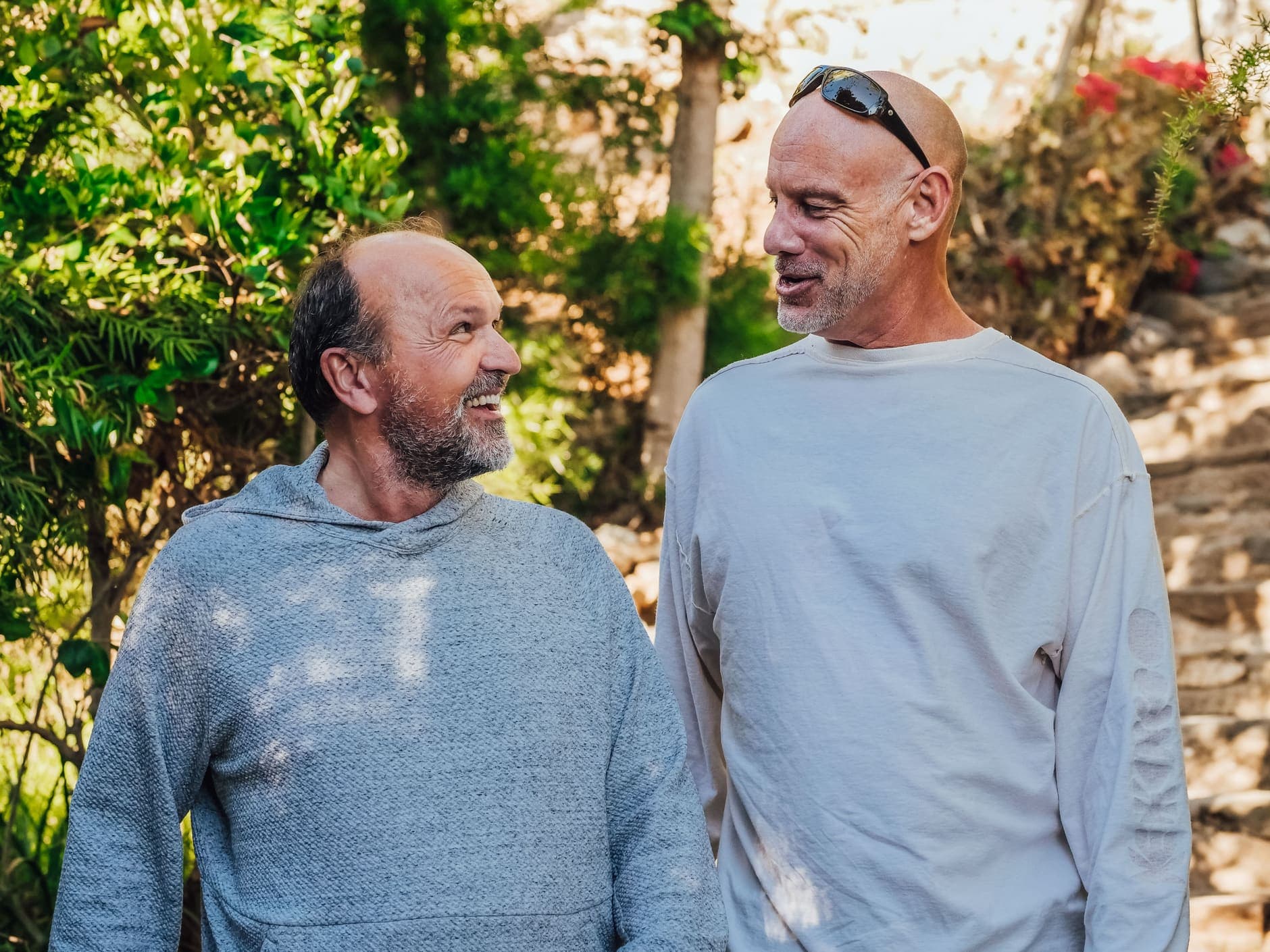 Dois homens de barba grisalha sorriem um para o outro ao ar livre, perto de plantas.