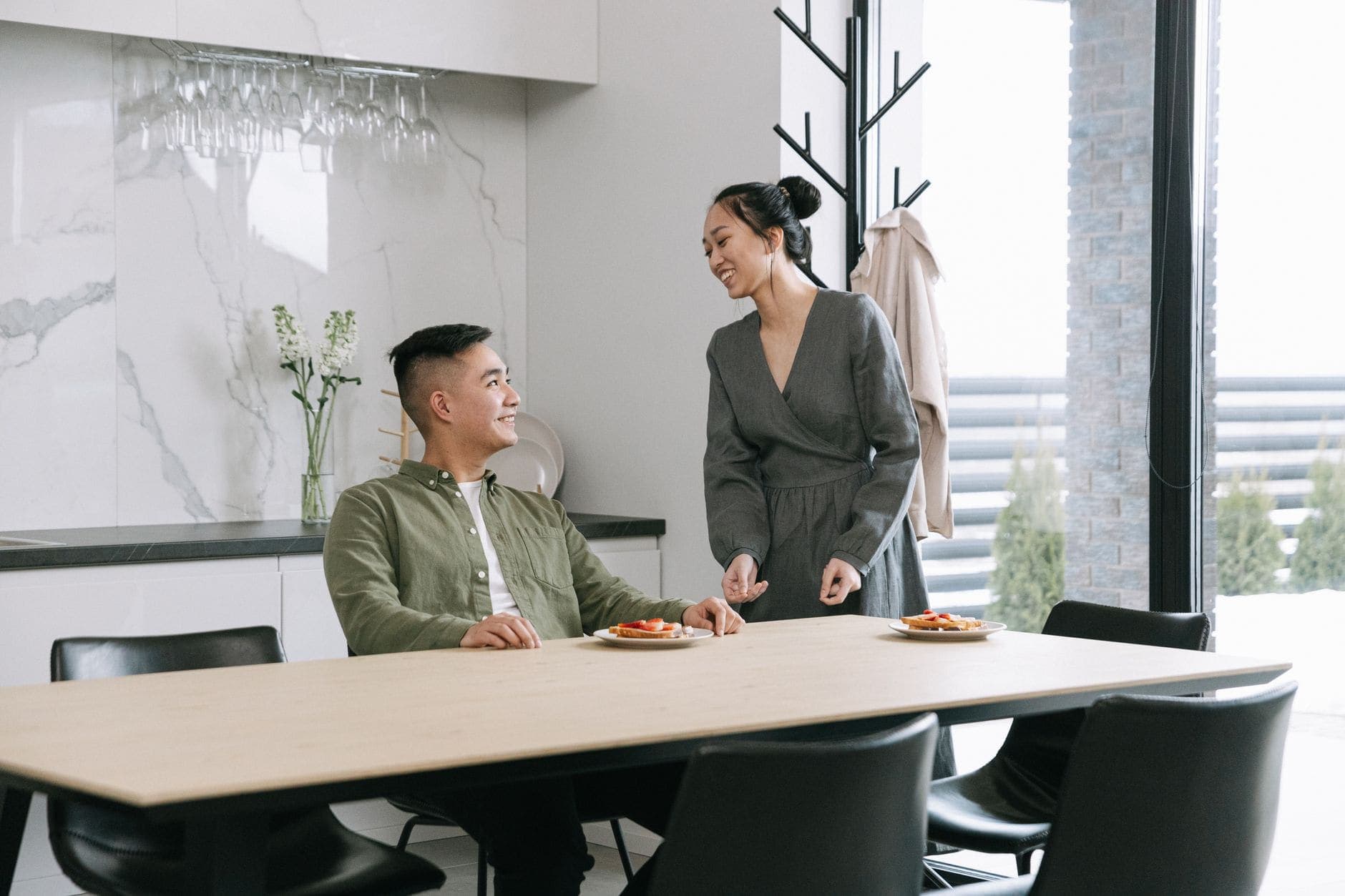Um homem sentado e uma mulher em pé sorriem um para o outro junto a uma mesa com pratos.