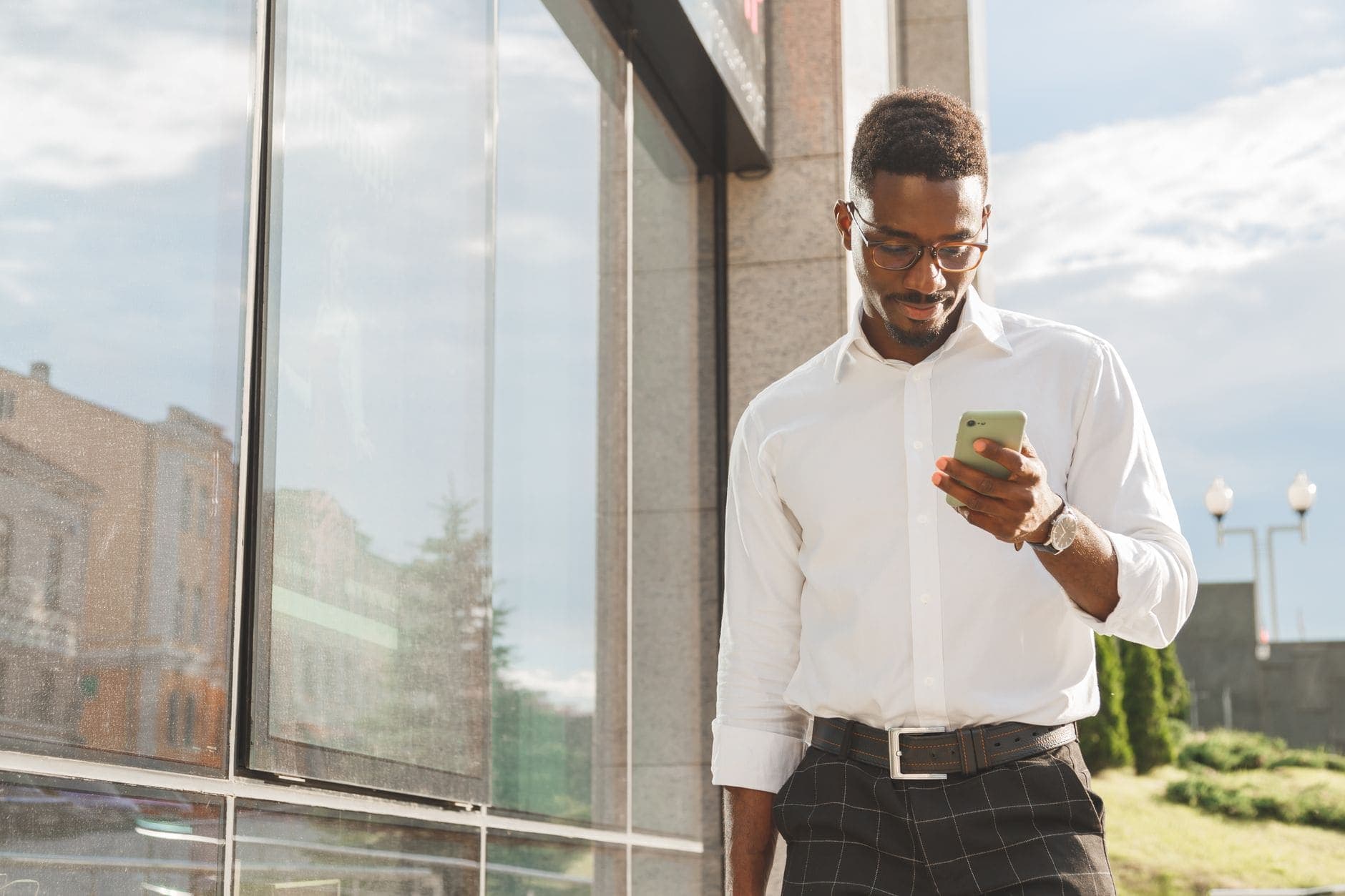 Um homem de camisa branca olha para o celular junto a uma fachada envidraçada.