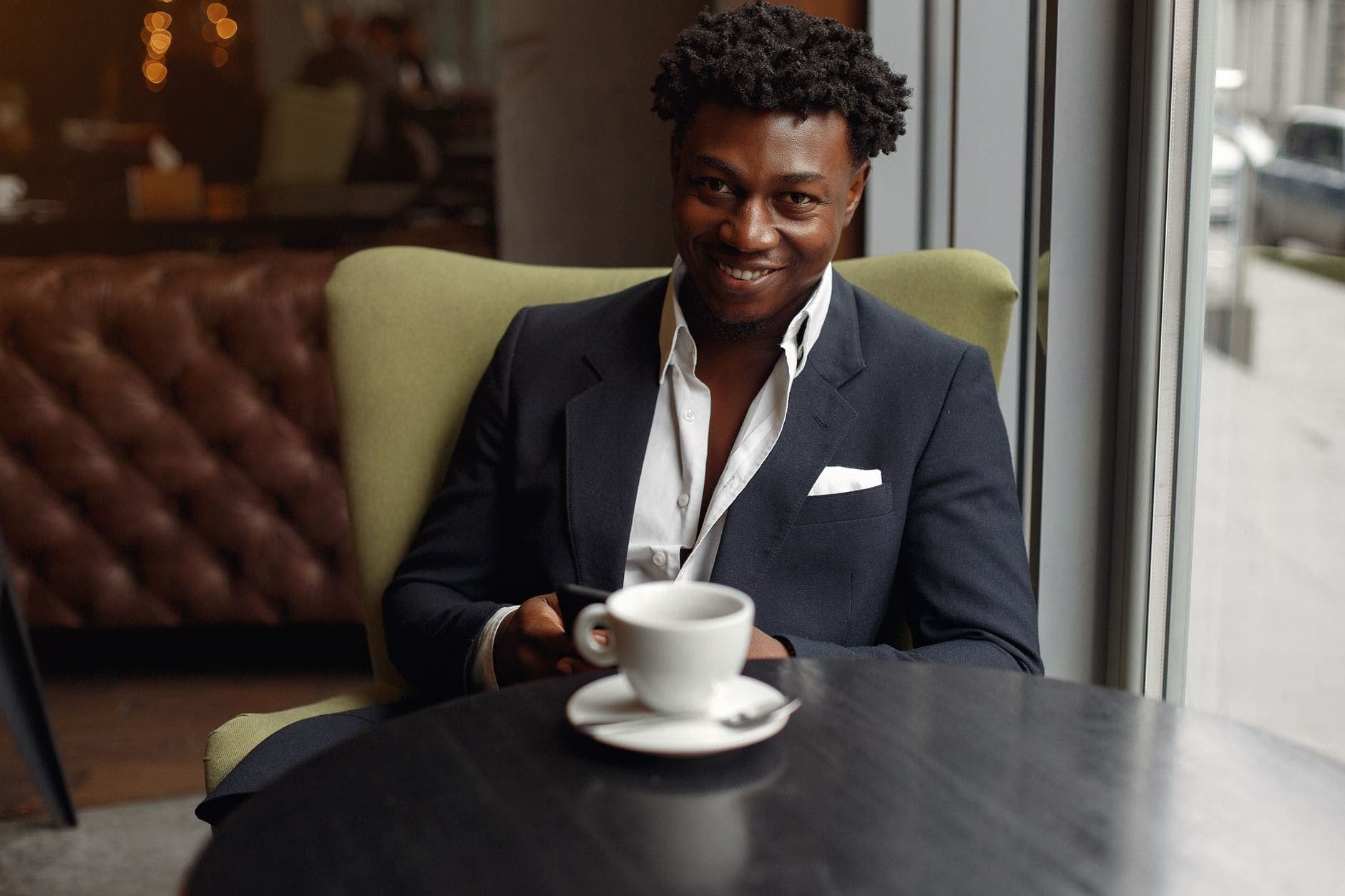 A man in a dark jacket smiles beside a café window, with a white cup on the table.