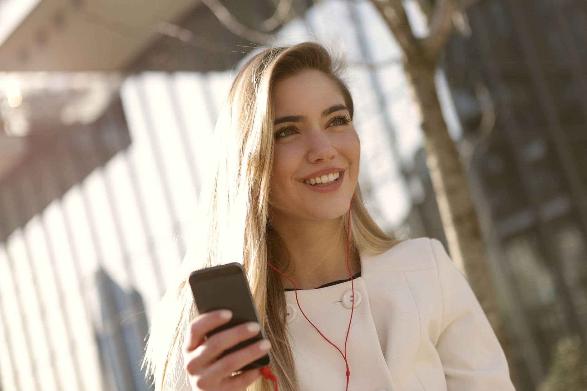 Eine lächelnde Frau im weissen Mantel hält ihr Smartphone und trägt rote Kopfhörer.