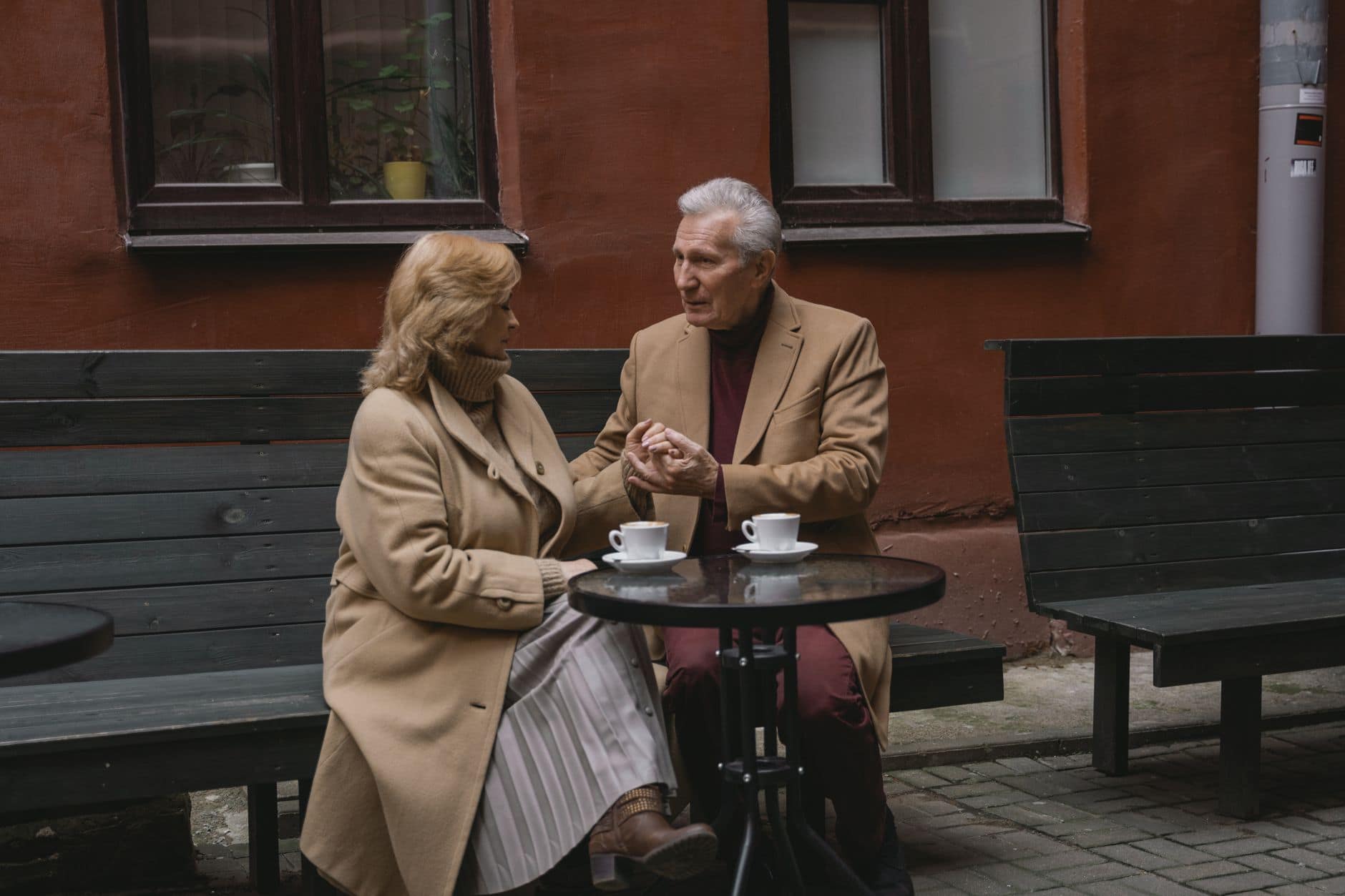 Ein Paar in beigen Mänteln unterhält sich bei Kaffee an einem kleinen Tisch im Freien.