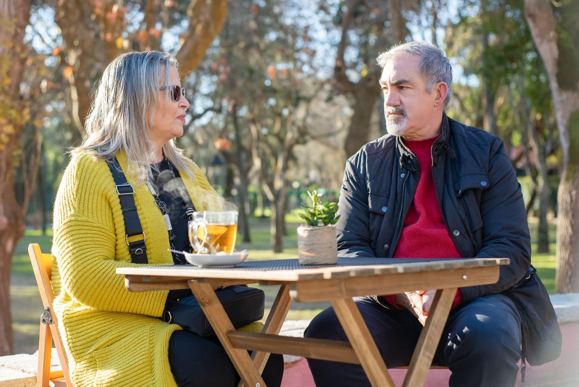 Mulher e homem maduros conversam sentados a uma mesa ao ar livre em um parque.