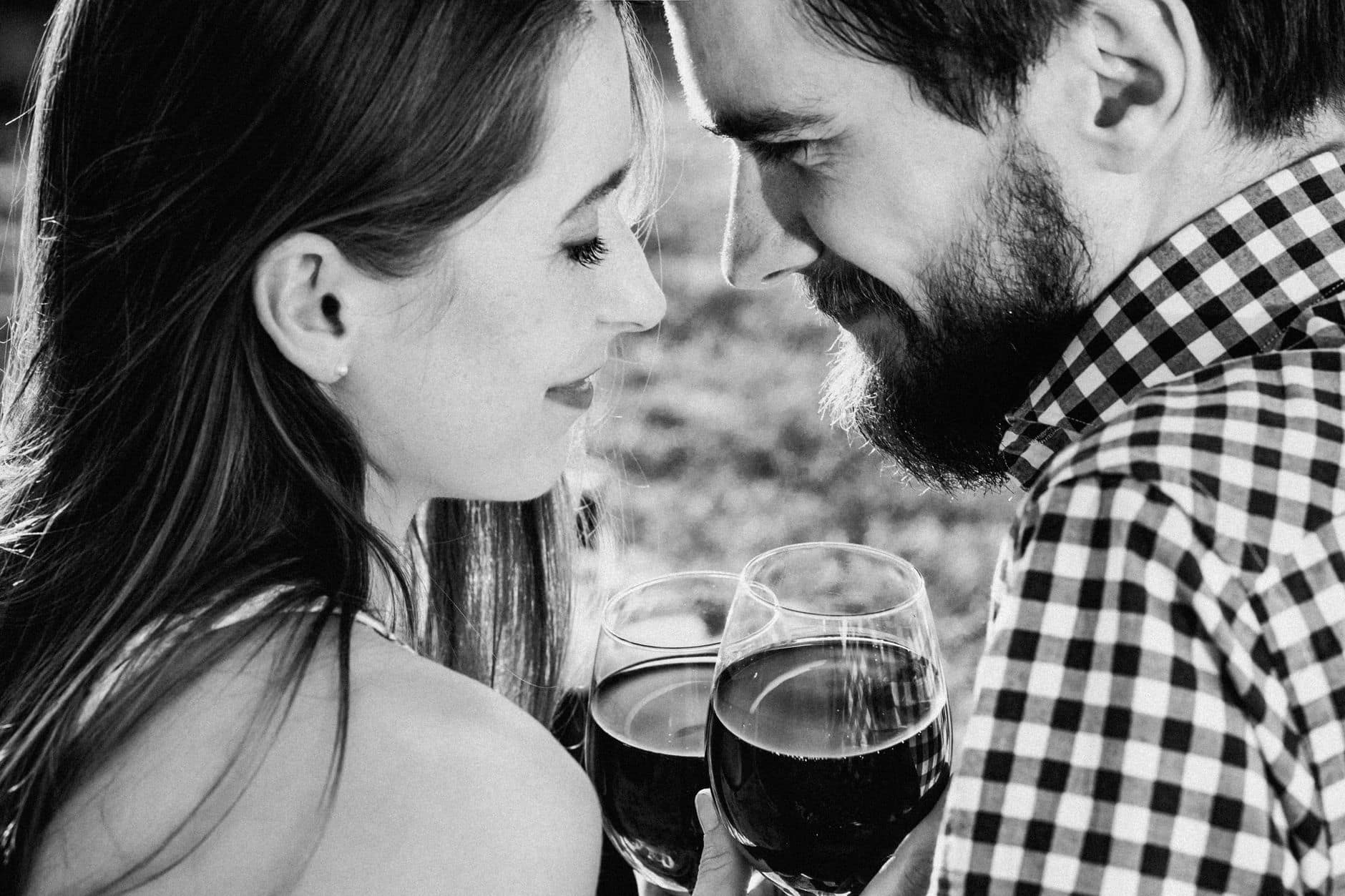 Fotografie alb-negru cu un cuplu care apropie frunțile, ținând pahare de vin