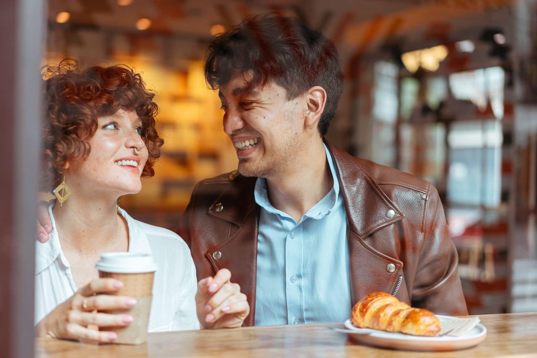 Een vrouw en een man lachen naar elkaar aan een cafétafel met koffie en een croissant.