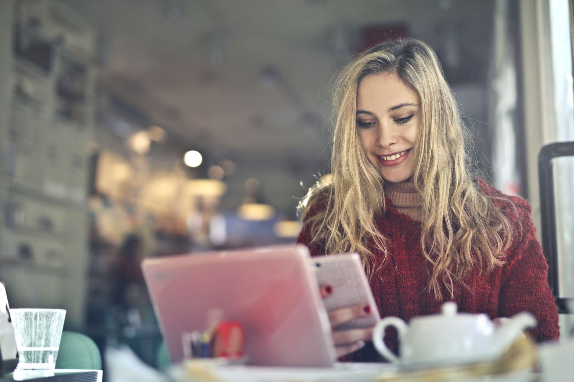 Une femme en pull bordeaux sourit devant un ordinateur portable et tient son téléphone.